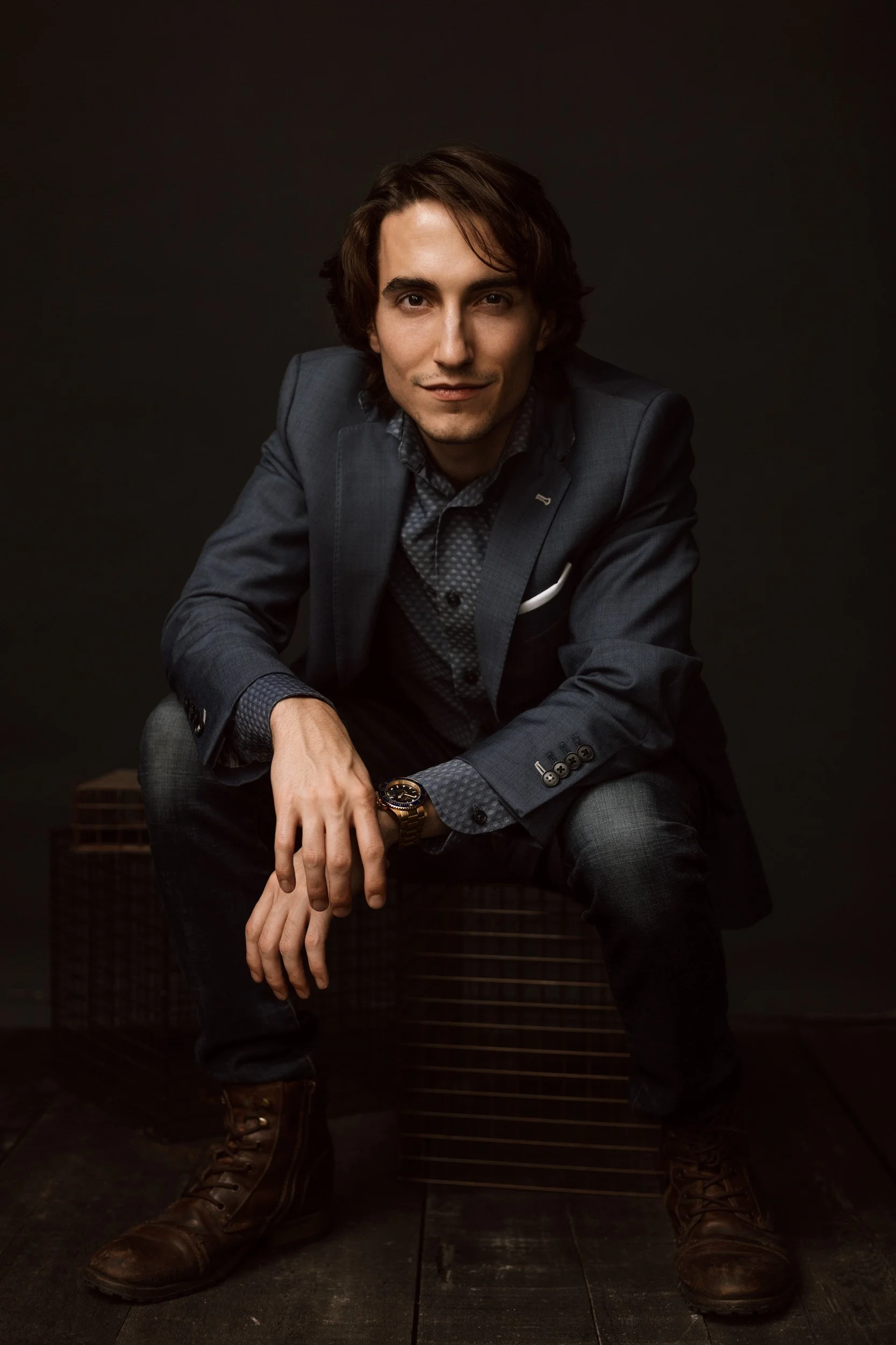 Moody studio portrait of a seated man in a dark suit leaning forward against a deep brown background.