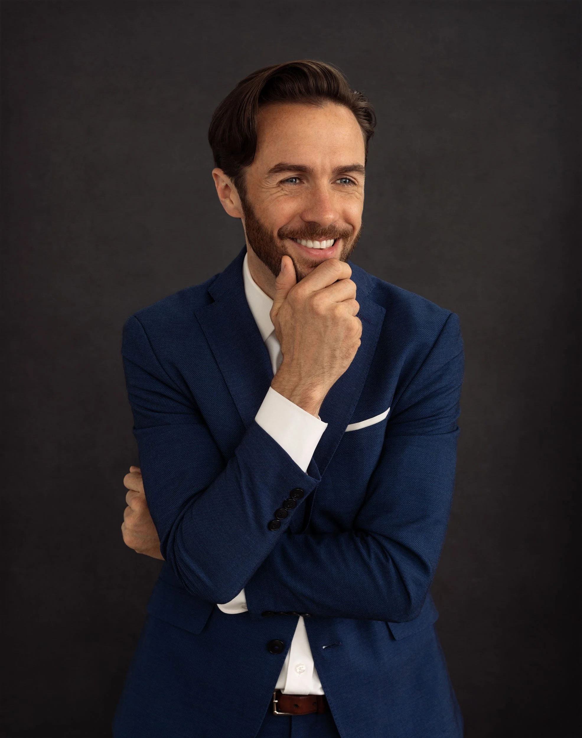 Professional studio portrait of a smiling man in a blue suit posed with one hand at his chin against a dark background.