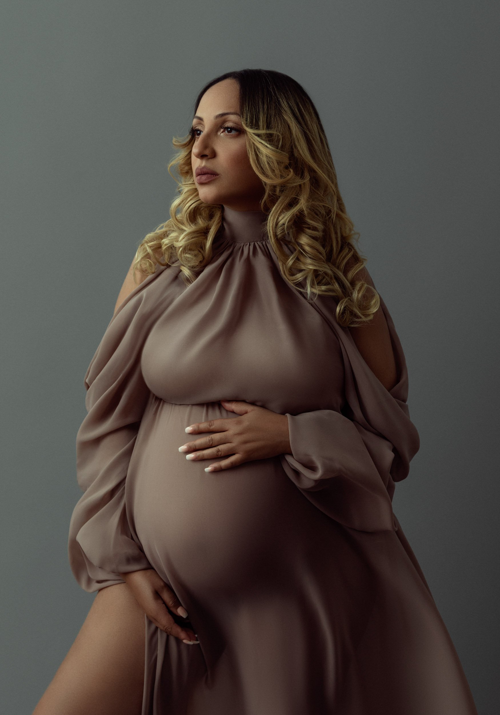 Studio maternity portrait of a pregnant woman wearing a flowing taupe dress against a soft gray background.