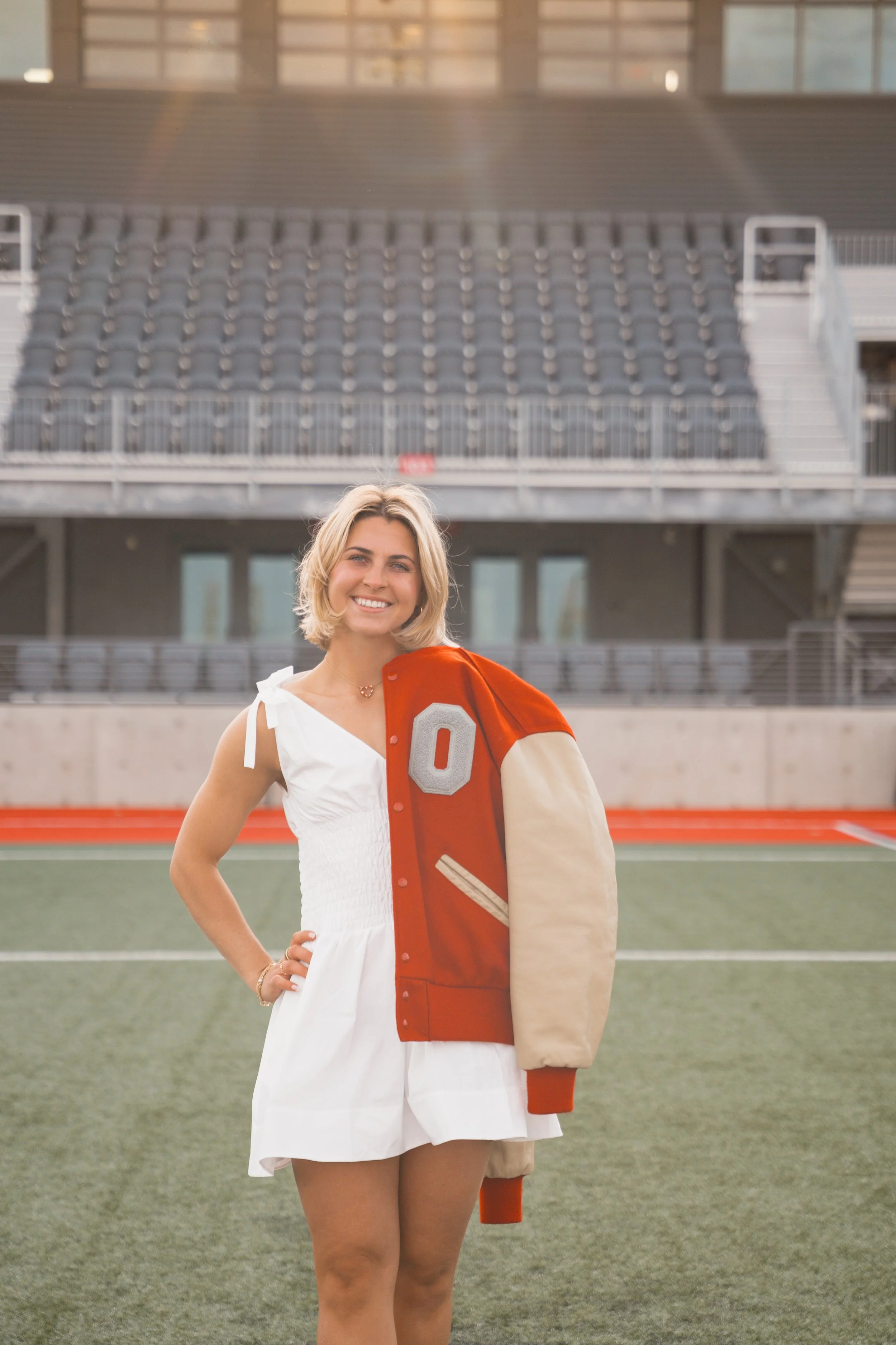 A woman standing on a football field holding a Ohio State varsity jacket with a Block "O" on it, smiling at the camera.