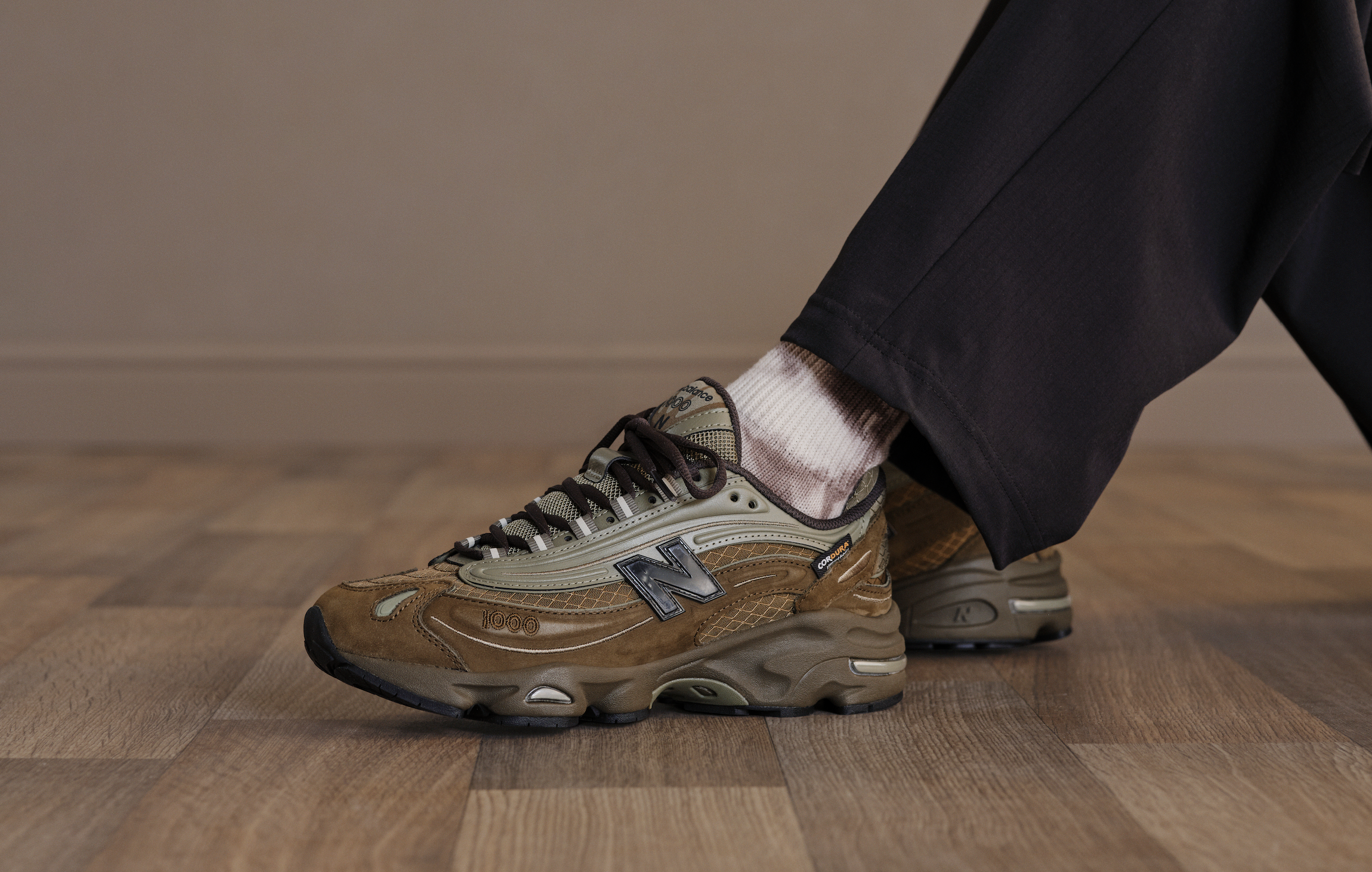 Close-up of a person wearing beige and brown New Balance sneakers, black pants, and pink socks on a wooden floor.