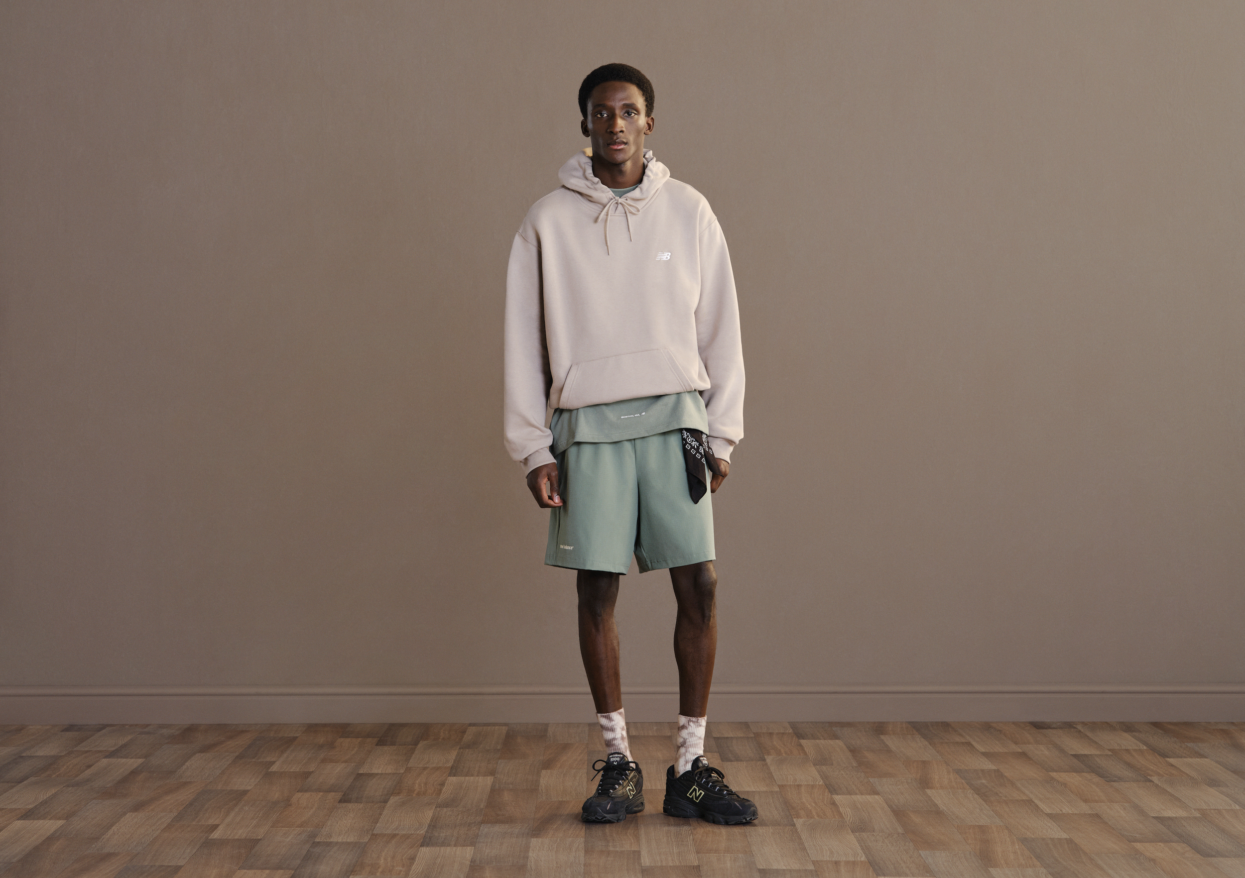 A young man with dark skin and short hair standing in an empty room with beige walls and a wooden floor. He is wearing a beige hoodie, loose mint green shorts, black sneakers, and patterned socks, holding a black bandana in his right hand.