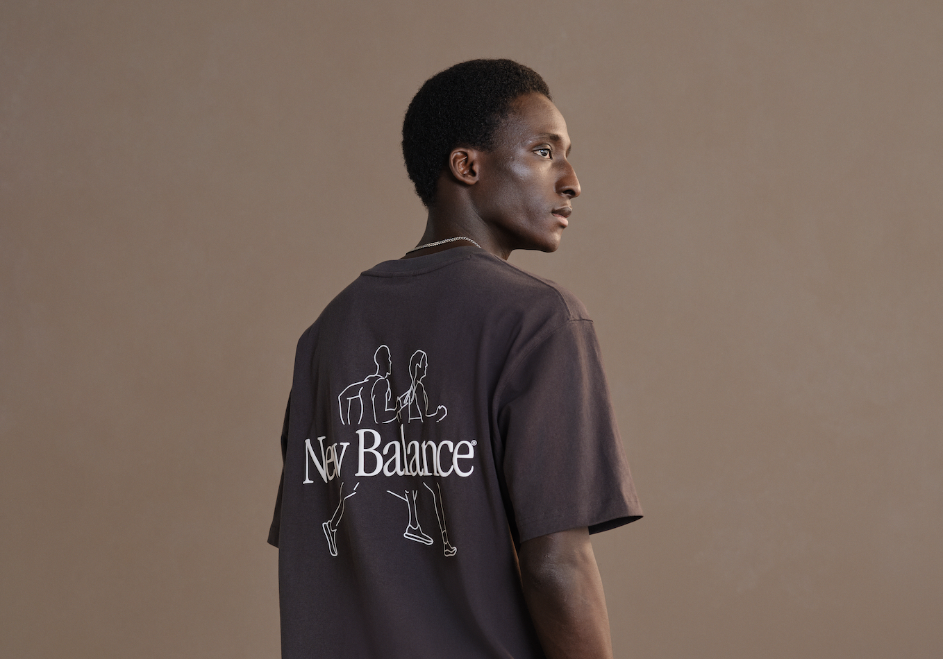 A young Black man with short curly hair, looking to the right, wearing a dark T-shirt with a New Balance logo and running graphic on the back, standing against a plain beige background.
