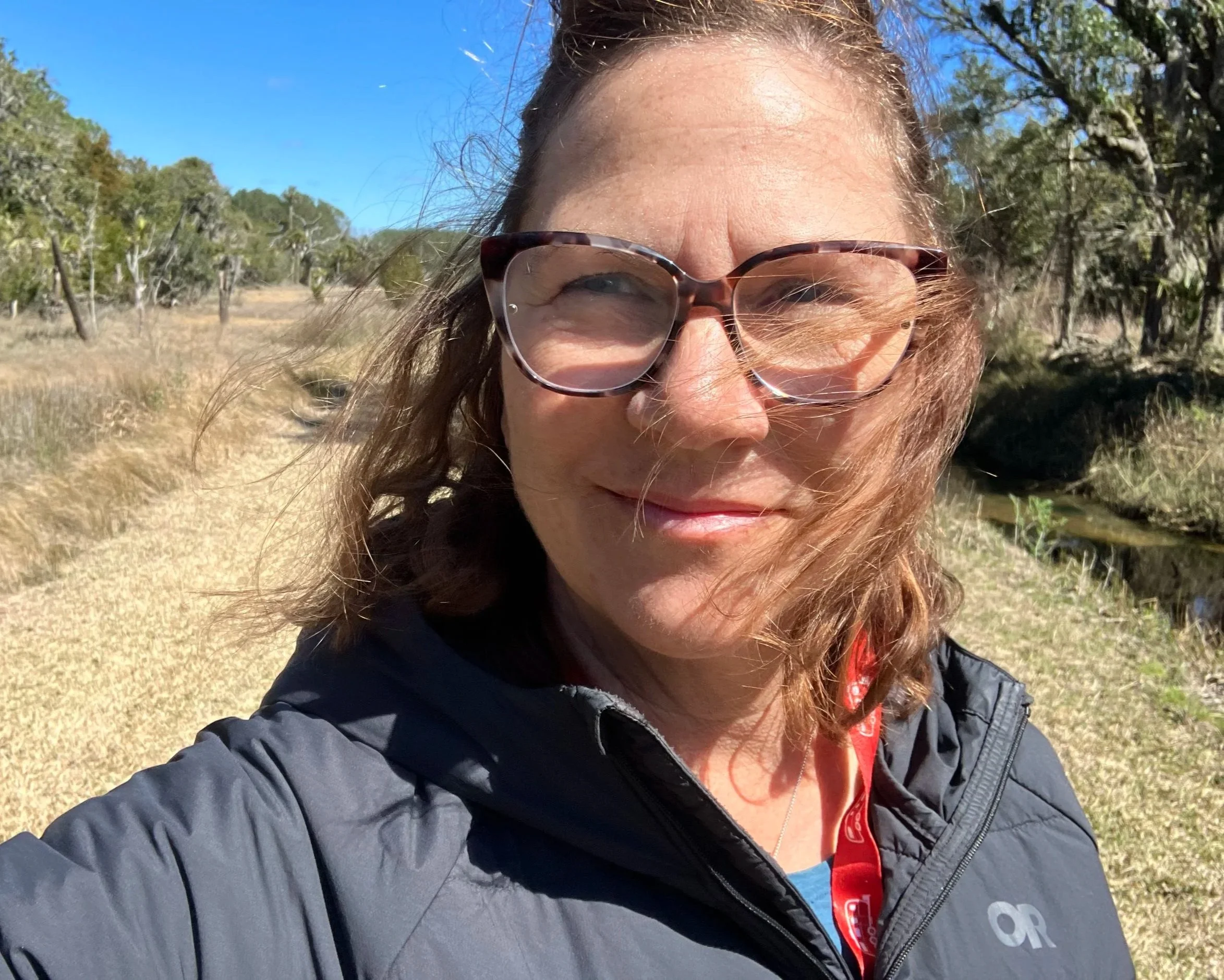 Caucasian woman with shoulder length brown hair and glasses wearing a jacket standing outside in a field on a sunny day