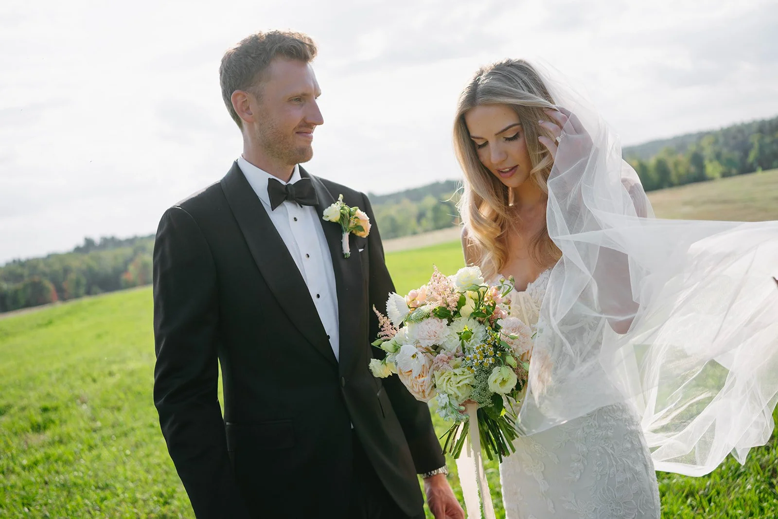 A bride and groom standing outdoors in a grassy field, with the bride holding a bouquet of flowers and the groom dressed in a black tuxedo with a bow tie, during a wedding ceremony or photo session.