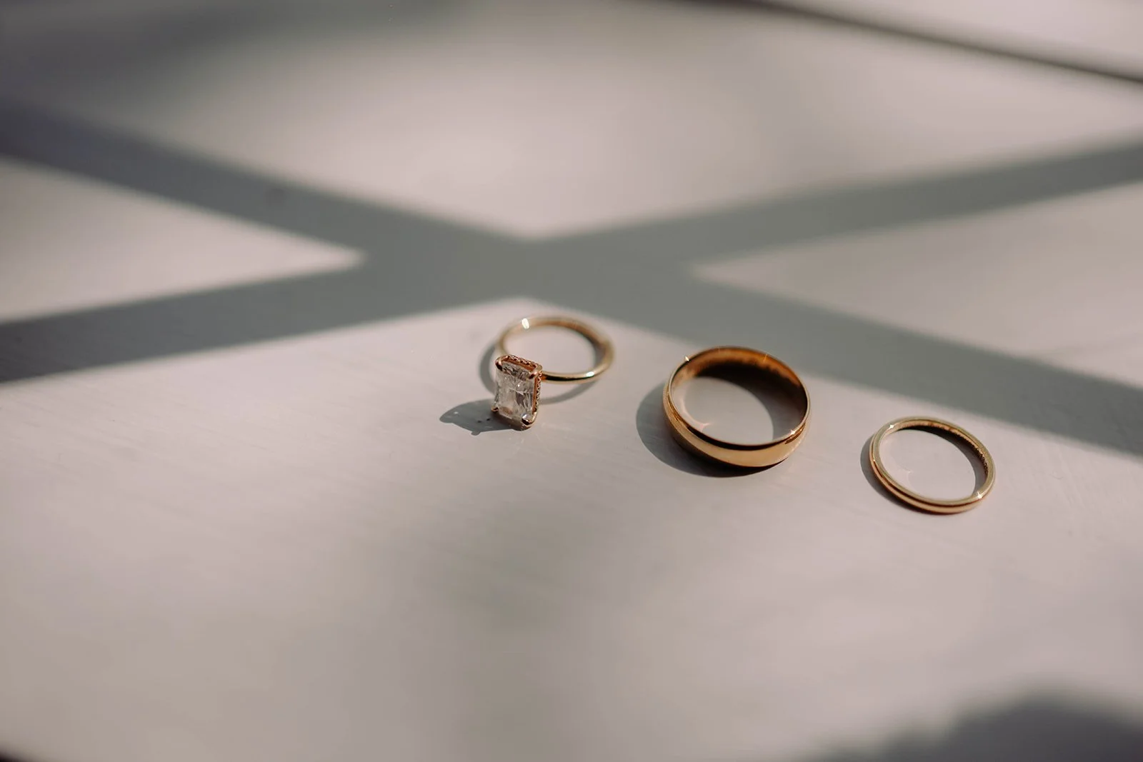 Three gold rings, one with a square-shaped gemstone, are arranged on a white surface with shadows from nearby blinds.