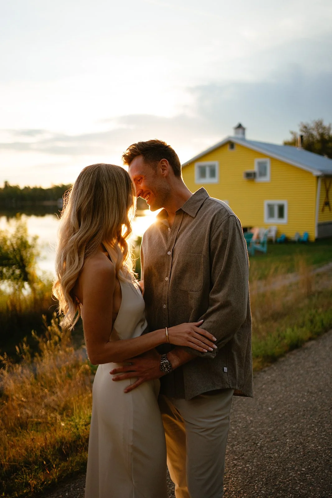 A couple smiling and embracing at sunset near a yellow house by a body of water.