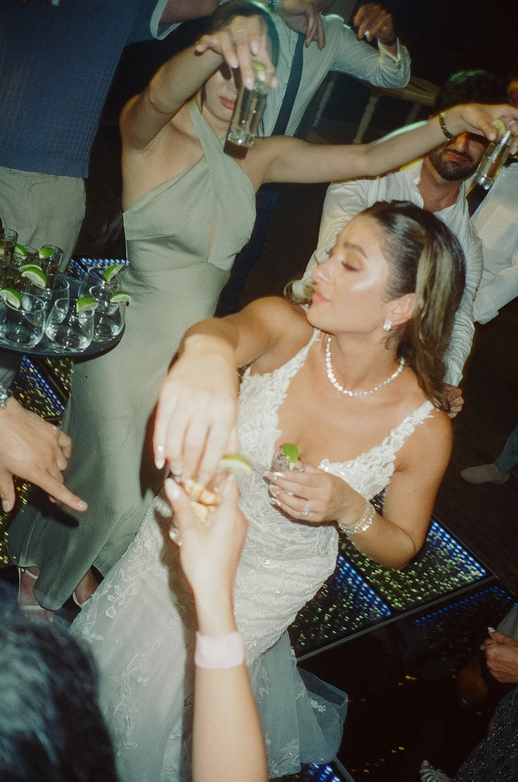 A woman in a wedding dress receiving a shot at a celebration or wedding reception, with others holding shot glasses and a tray of lime wedges nearby.