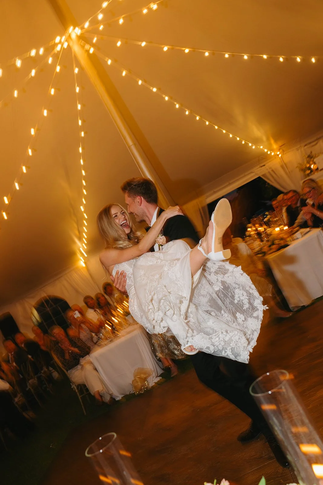 A bride and groom dancing together at a wedding reception under string lights inside a decorated tent, surrounded by seated guests.