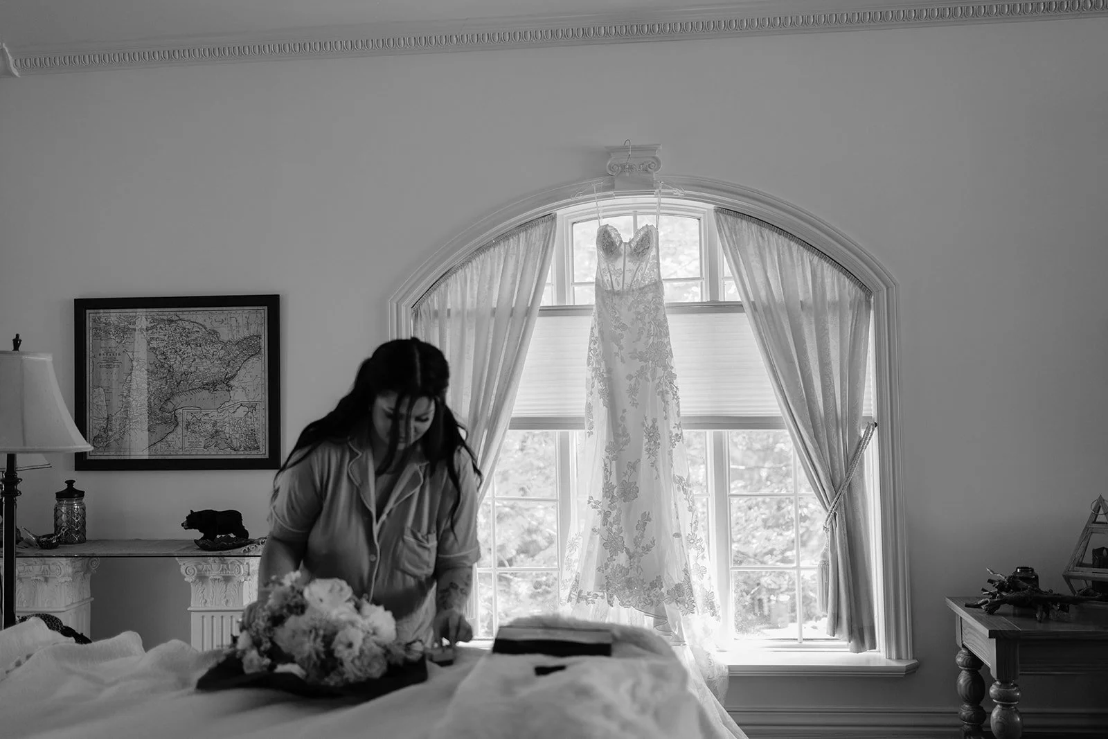 A woman with dark hair standing in a room, arranging a bouquet of flowers on a bed. In the background, a wedding dress hangs in front of a window with curtains. The room has a side table with a lamp and decorative items, and a framed map on the wall.
