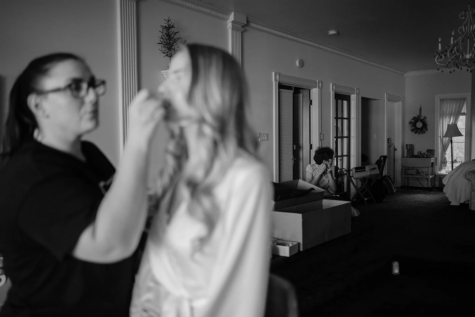 Black and white photo of people inside a house. Two women are in the foreground, one with glasses, and one with long hair, facing each other. In the background, a woman is sitting on a chair, covering her face with a tissue or cloth near a doorway.