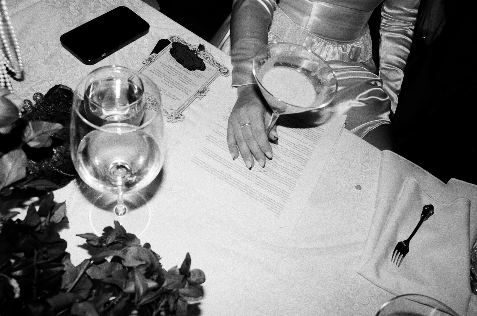 A woman wearing a satin robe at a table with a menu, a smartphone, empty wine glasses, a dessert cocktail, and a white napkin with a fork.