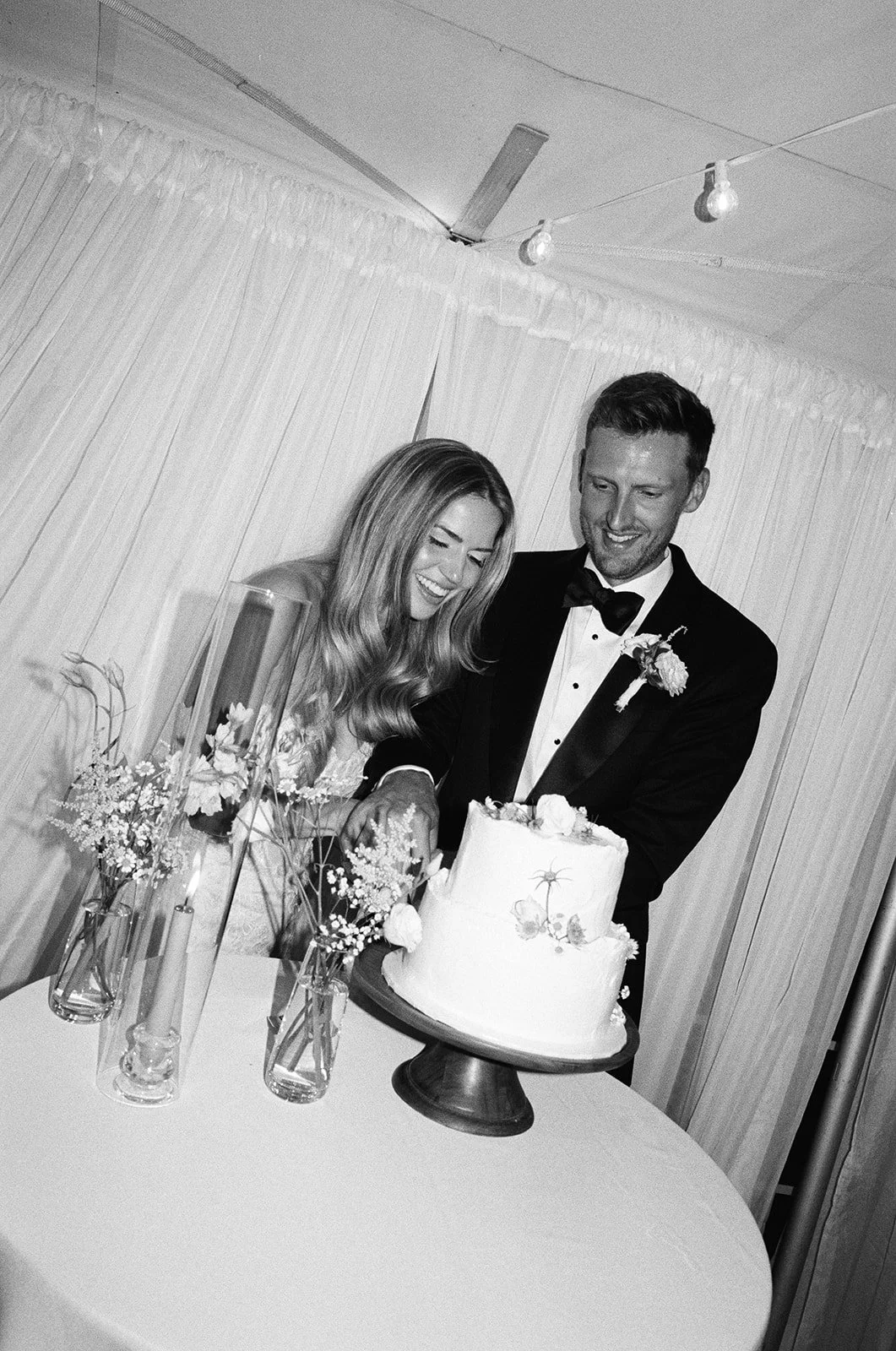 A black-and-white photo of a bride and groom cutting a wedding cake together at their wedding reception, with floral arrangements and candles on the table.
