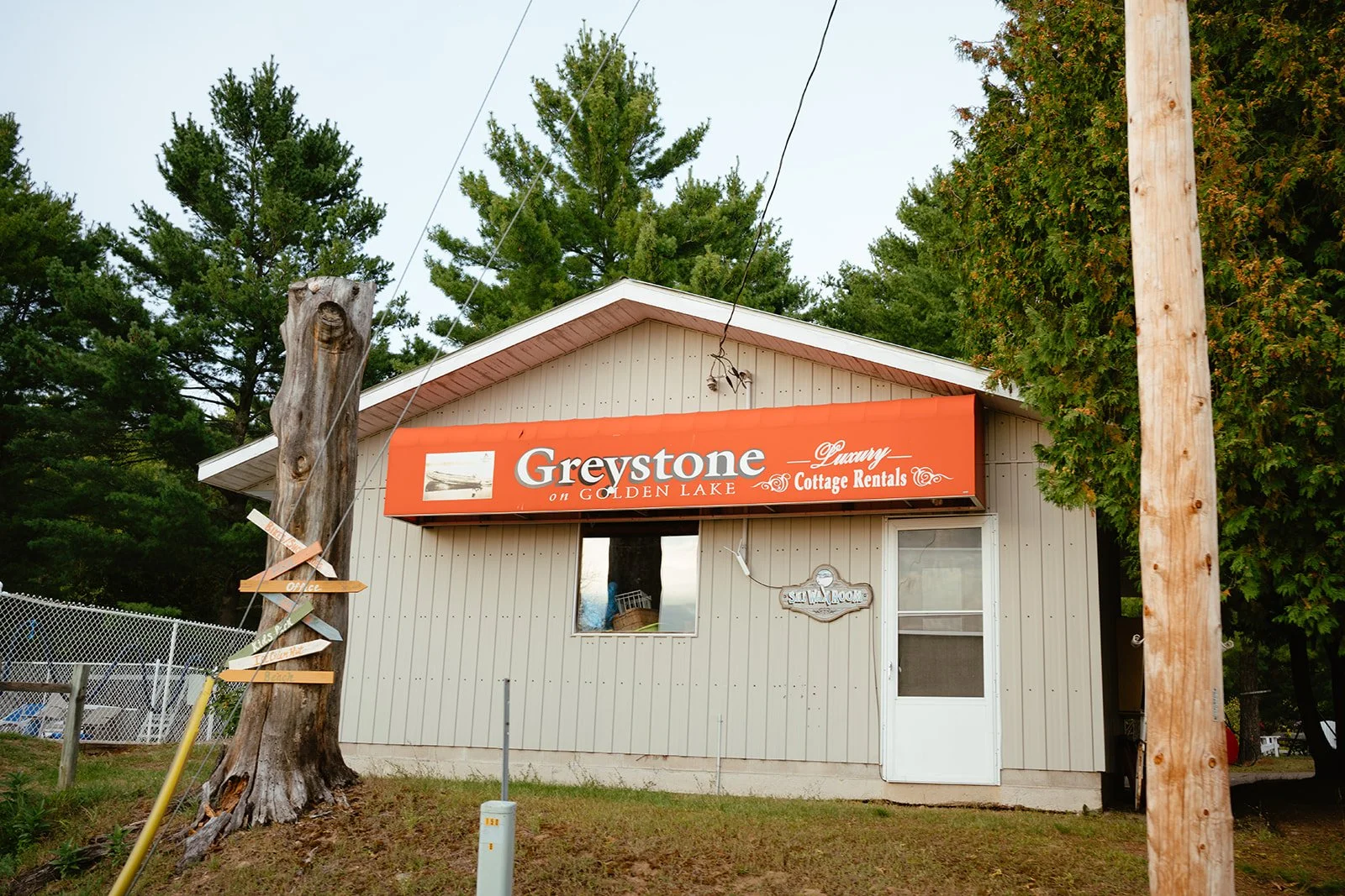 Small beige building with a red sign that reads "Greystone on Golden Lake, Luxury Cottage Rentals." Behind the building, tall green trees are visible, and a telephone pole with wires stands on the right. A weathered tree trunk with directional signs 