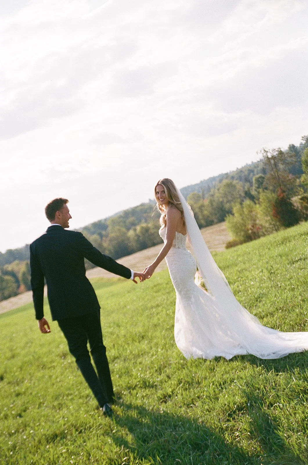 A bride and groom holding hands and walking across a grassy field on their wedding day, with trees and hills in the background.