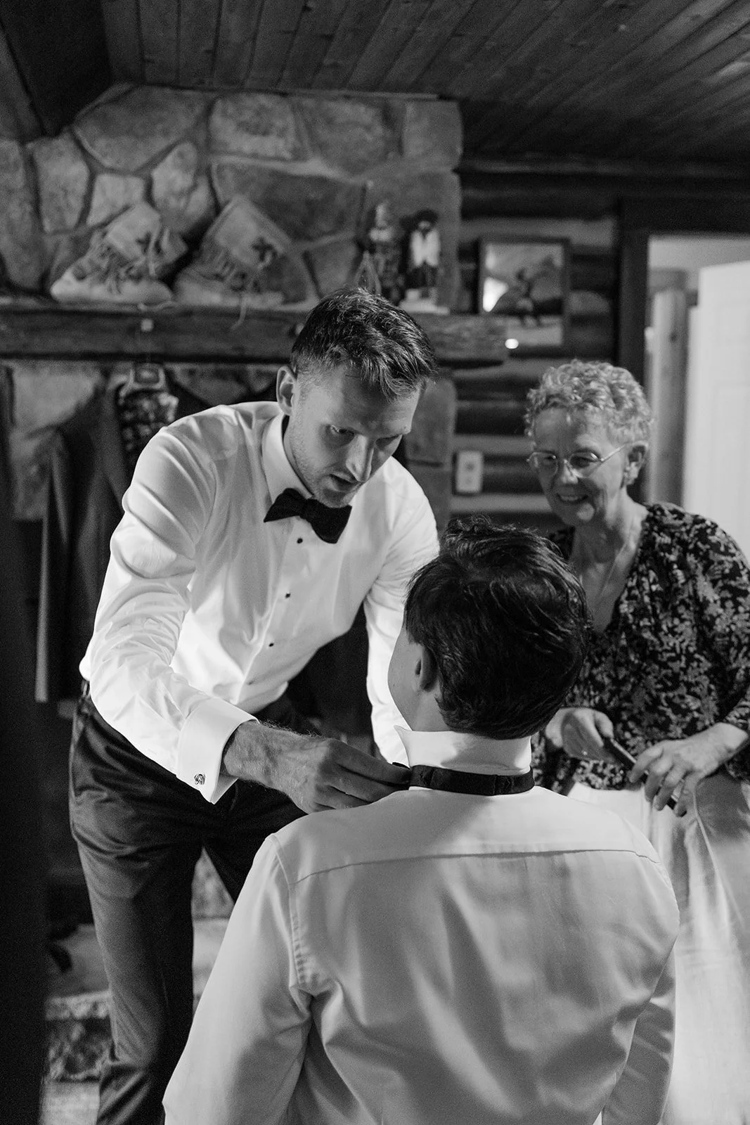 A young man in a tuxedo with a bow tie helps a young man in a white shirt put on a black bow tie while an older woman with curly hair and glasses watches smiling in a cozy log cabin with a stone fireplace in the background.