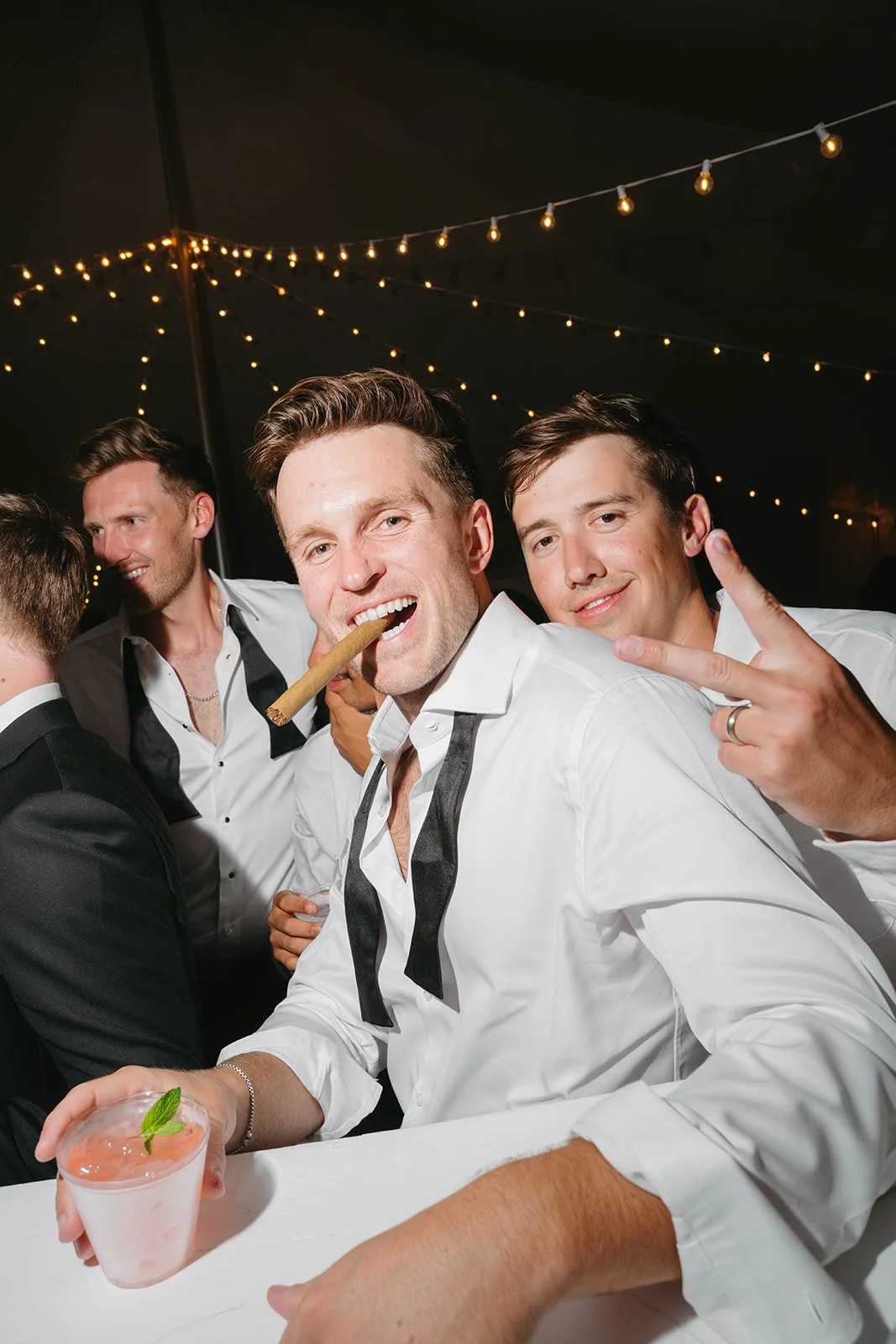 Group of men at a celebration or party, dressed in white shirts with black ties, under string lights, one man with a cigar in his mouth and another flashing a peace sign.