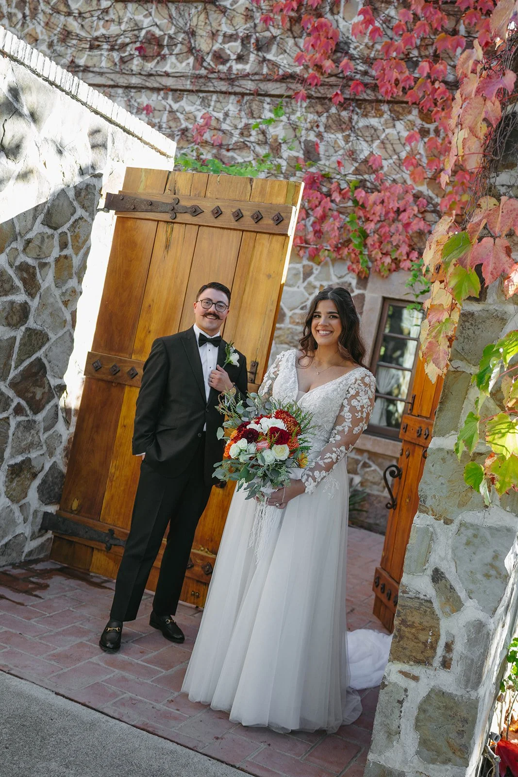 A bride and groom standing together outdoors in front of a stone wall with a wooden gate and autumnal foliage. The bride is holding a bouquet of red and white flowers, and the groom is wearing a black tuxedo with a bowtie and boutonniere.