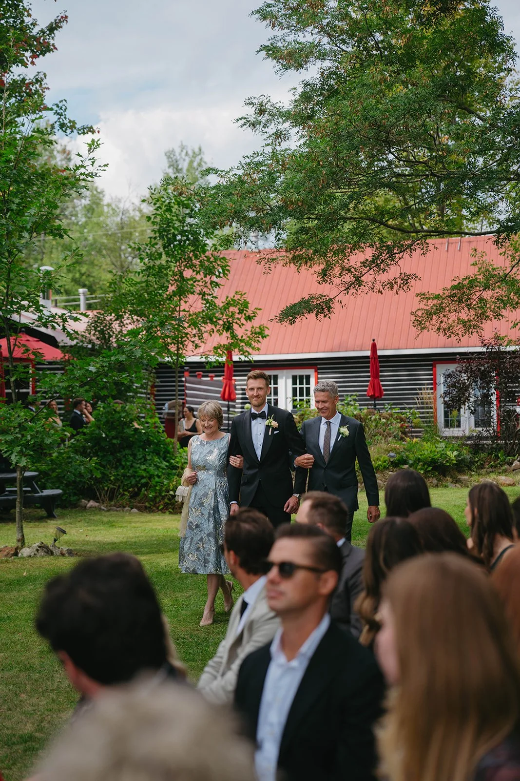 A bride and groom walking down the aisle outdoors, accompanied by a woman in a floral dress, with guests seated and standing on lush green grass under a partly cloudy sky.