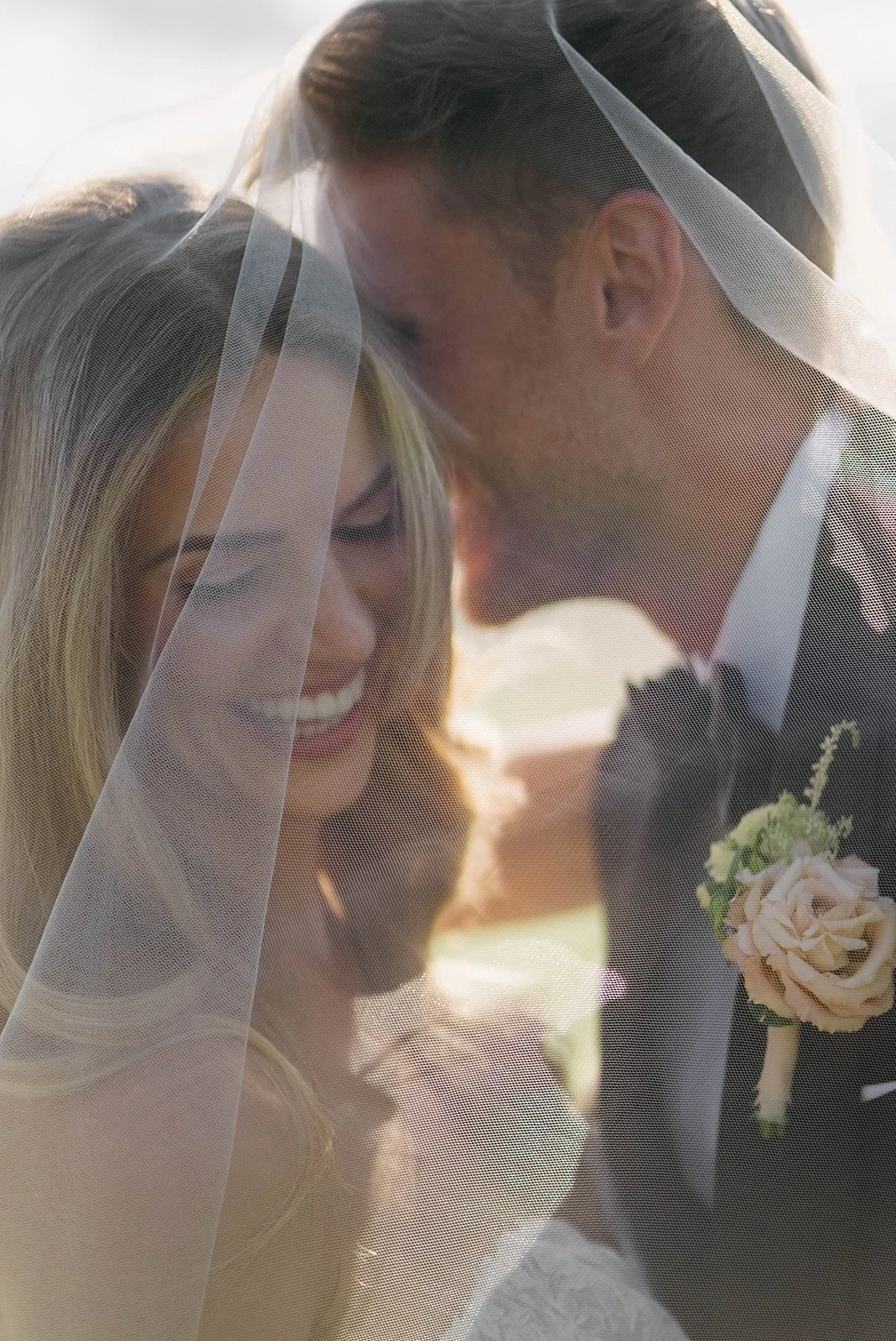 A bride and groom share an intimate moment under a veil with sunlight in the background.