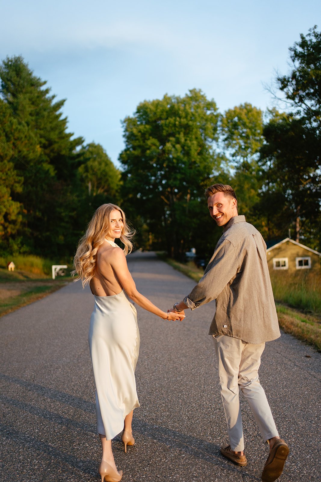 A smiling woman in a satin dress and heels holding hands with a smiling man in beige pants and a gray shirt on a rural road with trees and a small house in the background during sunset.