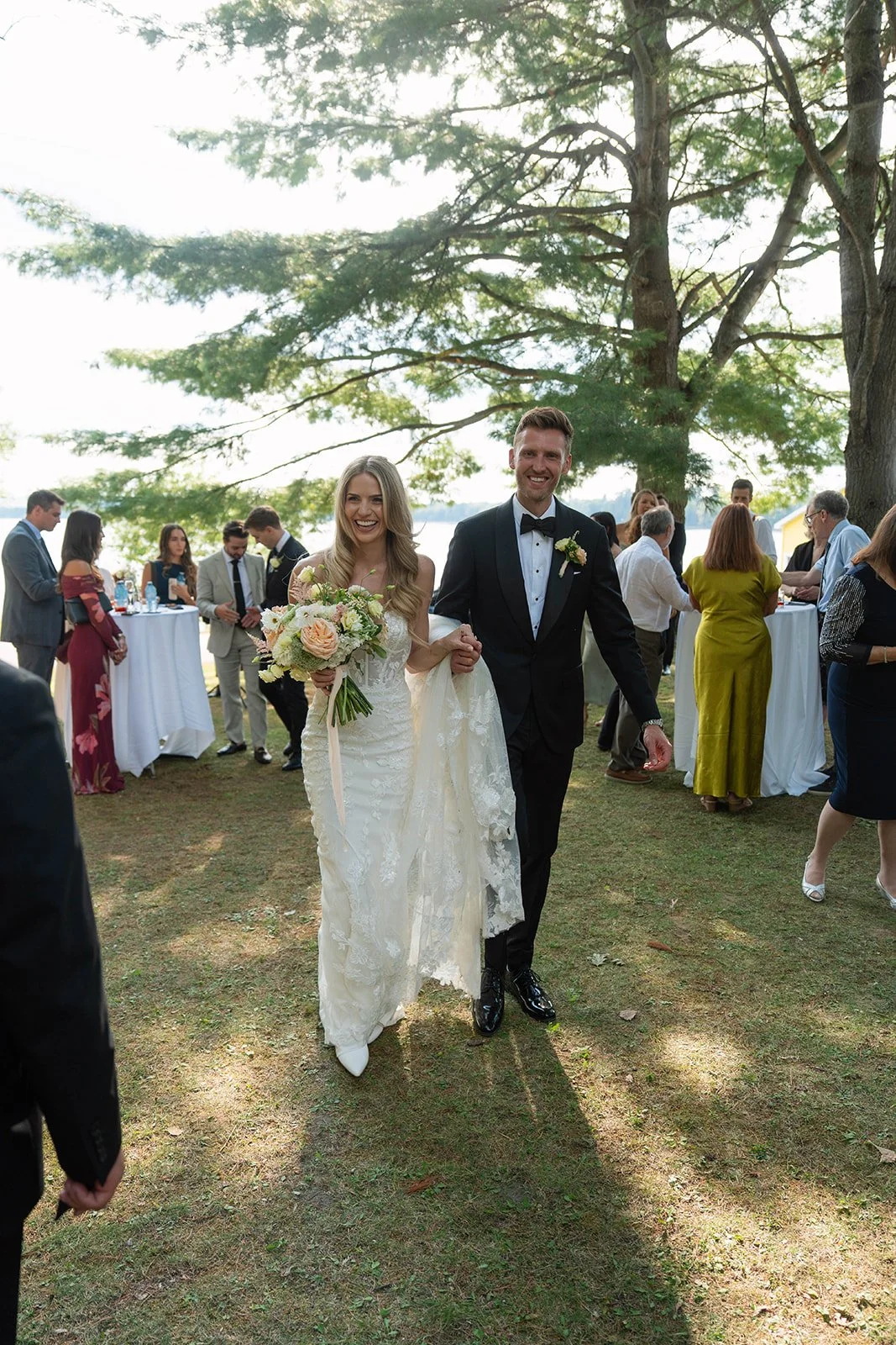 A newlywed couple walking arm-in-arm outdoors at their wedding reception, surrounded by guests under large trees, with a view of water in the background.