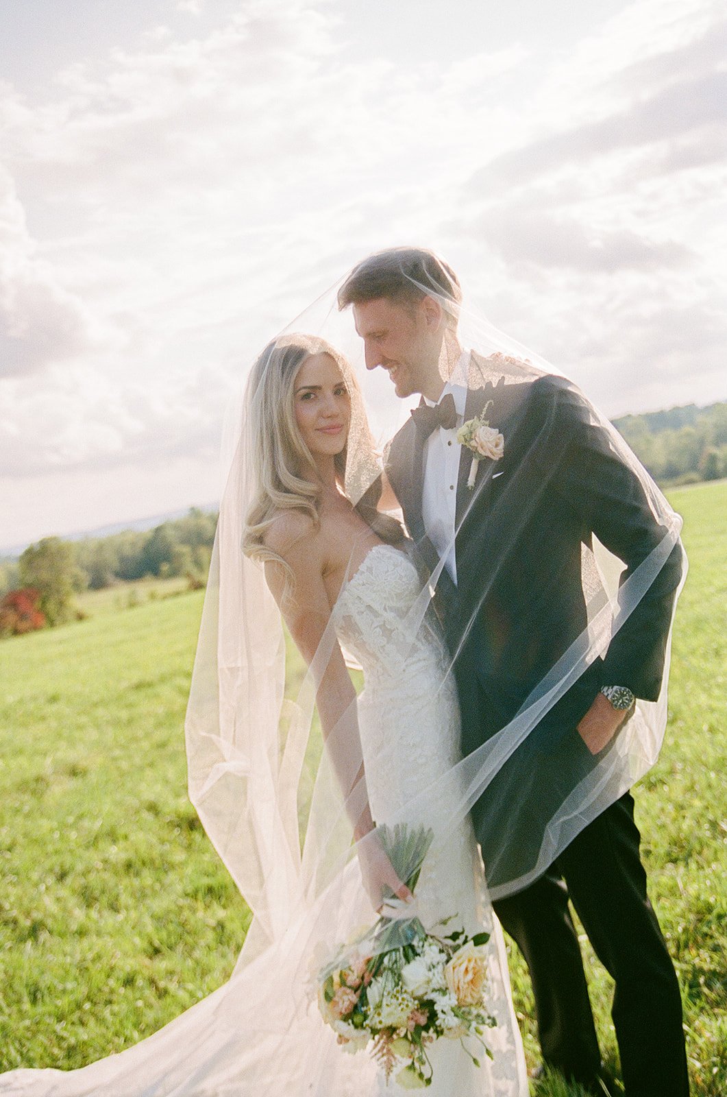 A bride and groom in wedding attire standing outdoors in a grassy field, sharing a moment under a veil with a scenic landscape and cloudy sky in the background.