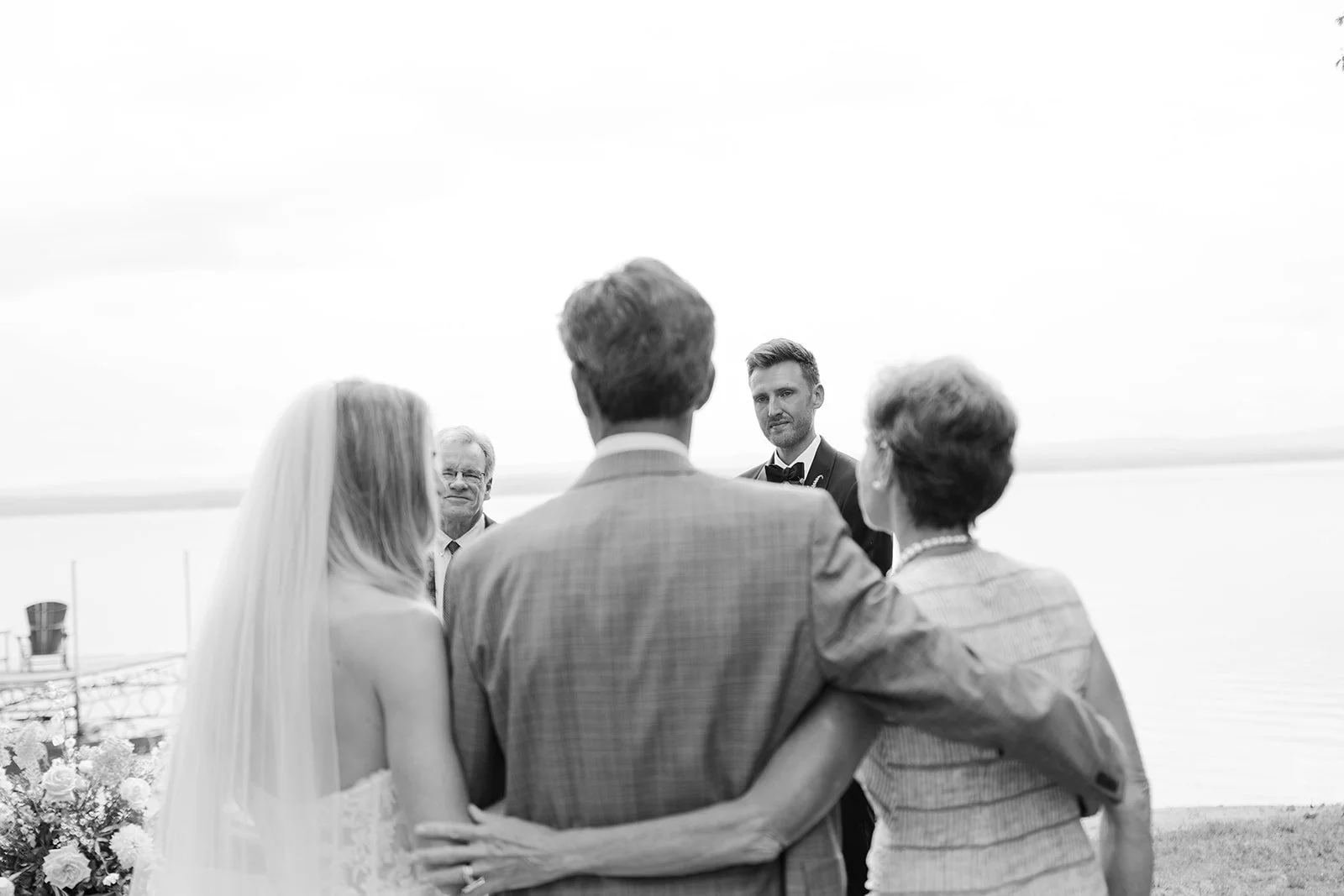 Black and white photo of a wedding ceremony by the water, with five people, including a bride in a wedding dress and veil, a groom in a suit, and three older adults, one of whom is officiating.