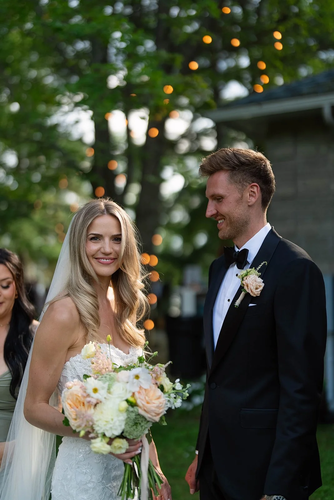 A bride and groom smiling and looking at each other during their outdoor wedding ceremony. The bride is holding a bouquet of pink, white, and peach flowers, and the groom is dressed in a black tuxedo with a white shirt and black bow tie. There are bl