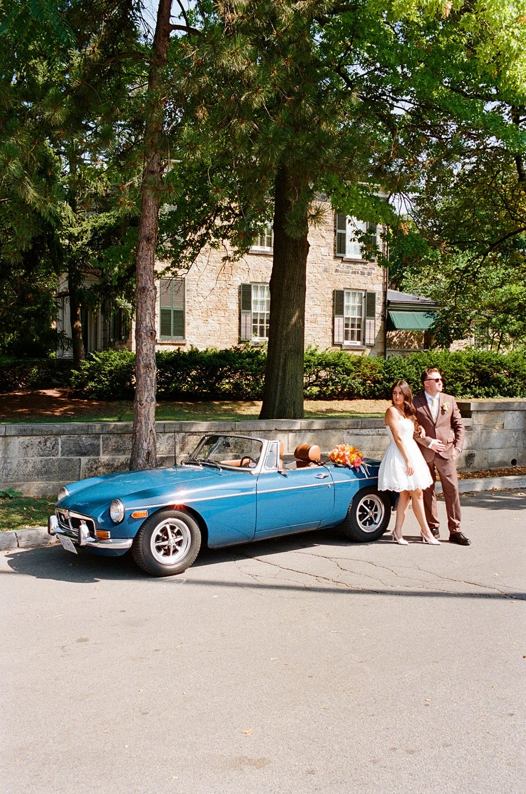 A bride and groom standing next to a blue vintage convertible car decorated with a bouquet of flowers, on a street lined with trees and a stone house.