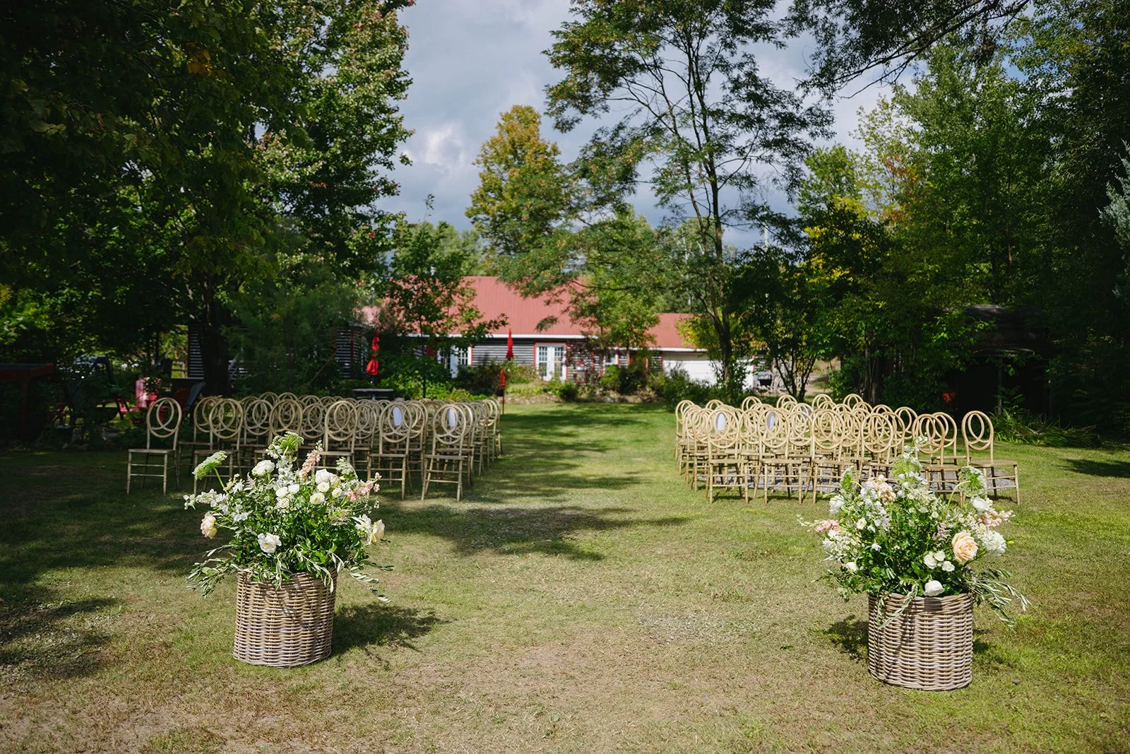 Outdoor wedding setup with rows of chairs and large flower arrangements on the grass, trees surrounding, and a building with a red roof in the background.
