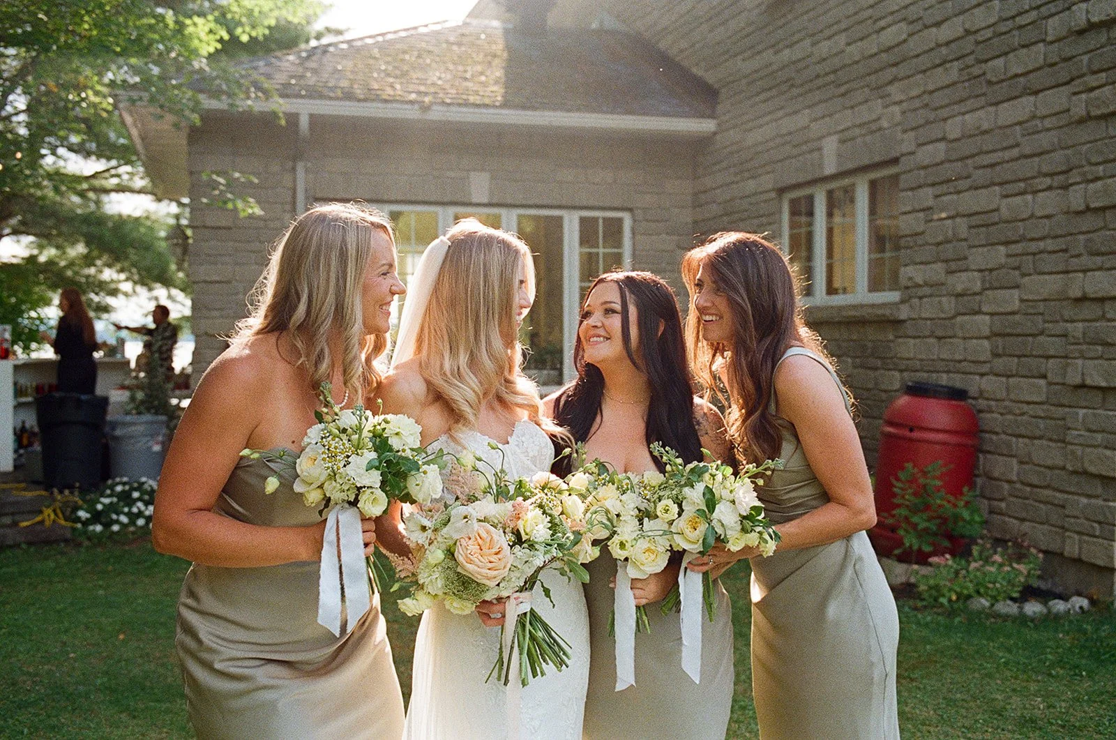Four women at a wedding, smiling and holding bouquets of white and peach flowers, standing outdoors near a brick house, with sunlight shining on them.