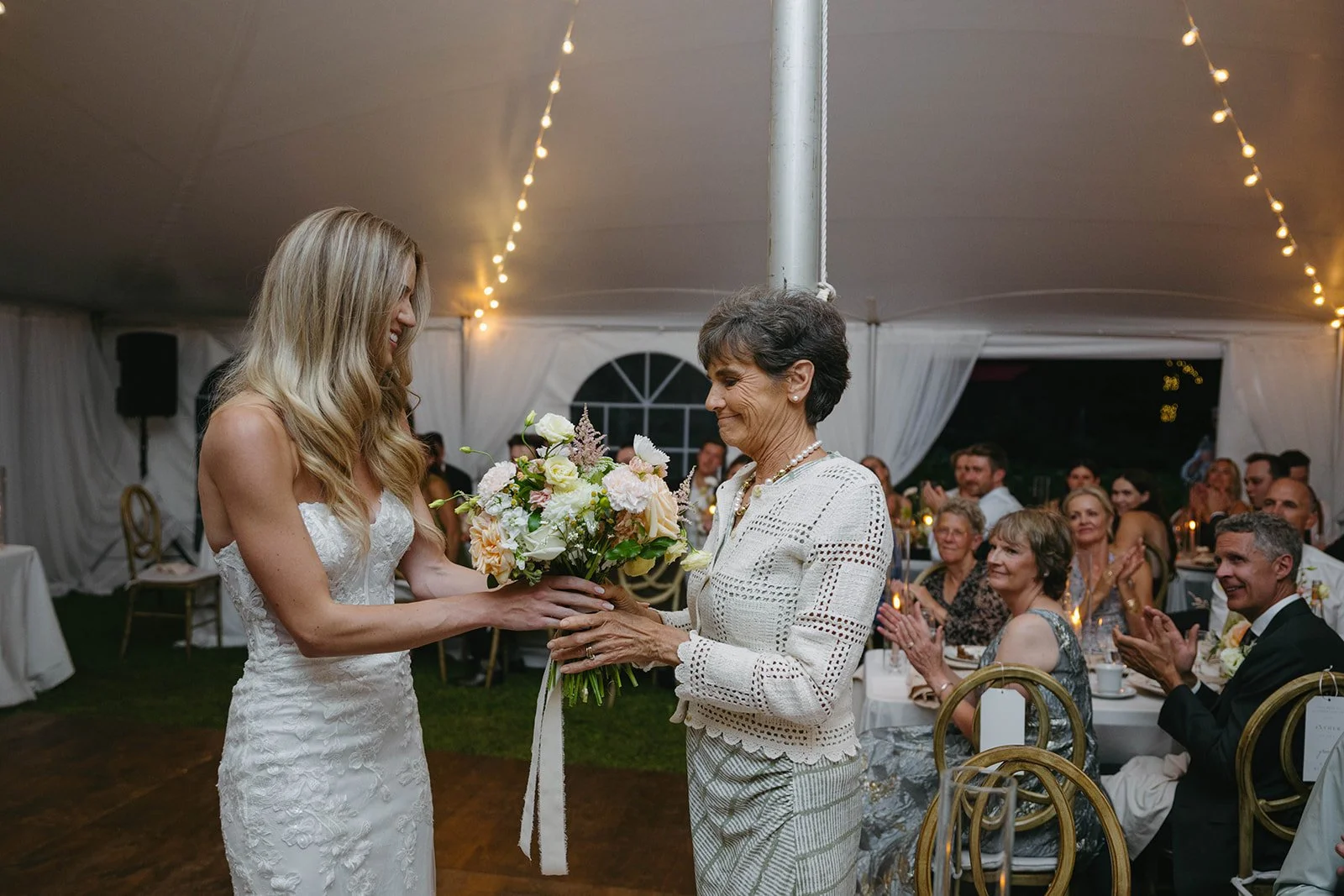 A bride in a white lace wedding dress receiving a bouquet of flowers from an older woman in a white crochet jacket at a wedding reception under a tent. Guests seated at tables in the background cheer and applaud.