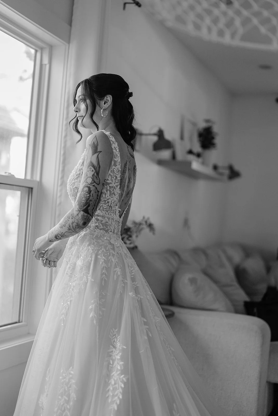 A bride in a lace wedding dress with tattoos on her arms stands near a window, looking outside, in a living room with a sofa and wall-mounted shelves.
