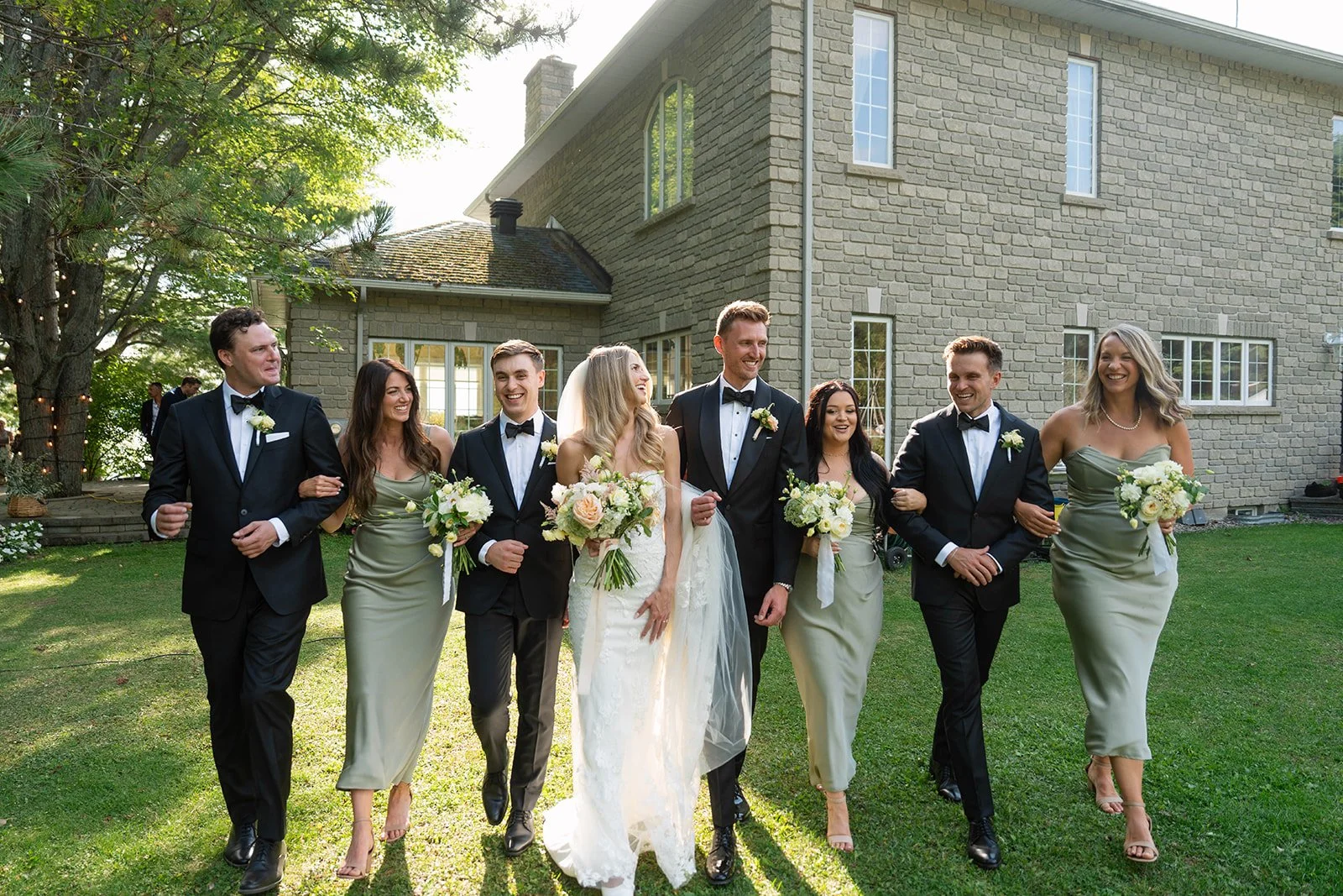A wedding party of eight walking arm-in-arm on a lush green lawn in front of a stone house, with the bride and groom in the center, holding bouquets, and bridesmaids and groomsmen on either side, all smiling and dressed in formal attire.