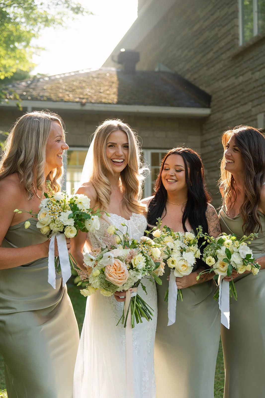 Four women, including a bride in a white dress, standing outdoors in sunlight with three bridesmaids in matching green dresses, all holding bouquets of white and peach flowers, smiling and celebrating.