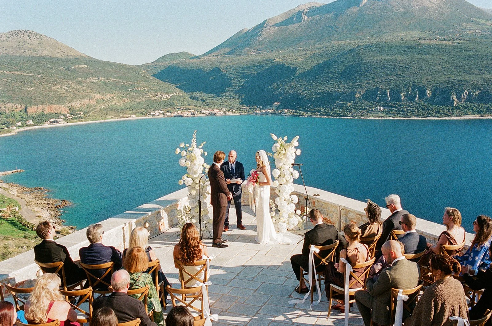 Outdoor wedding ceremony on a terrace overlooking a lake surrounded by green mountains, with a bride and groom exchanging vows under floral arches, and guests seated nearby.
