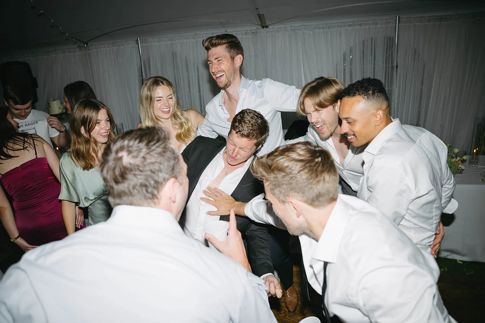 Group of people celebrating and laughing at a party, gathered around a man in a suit. They are indoors, with some women and men in semi-formal attire.
