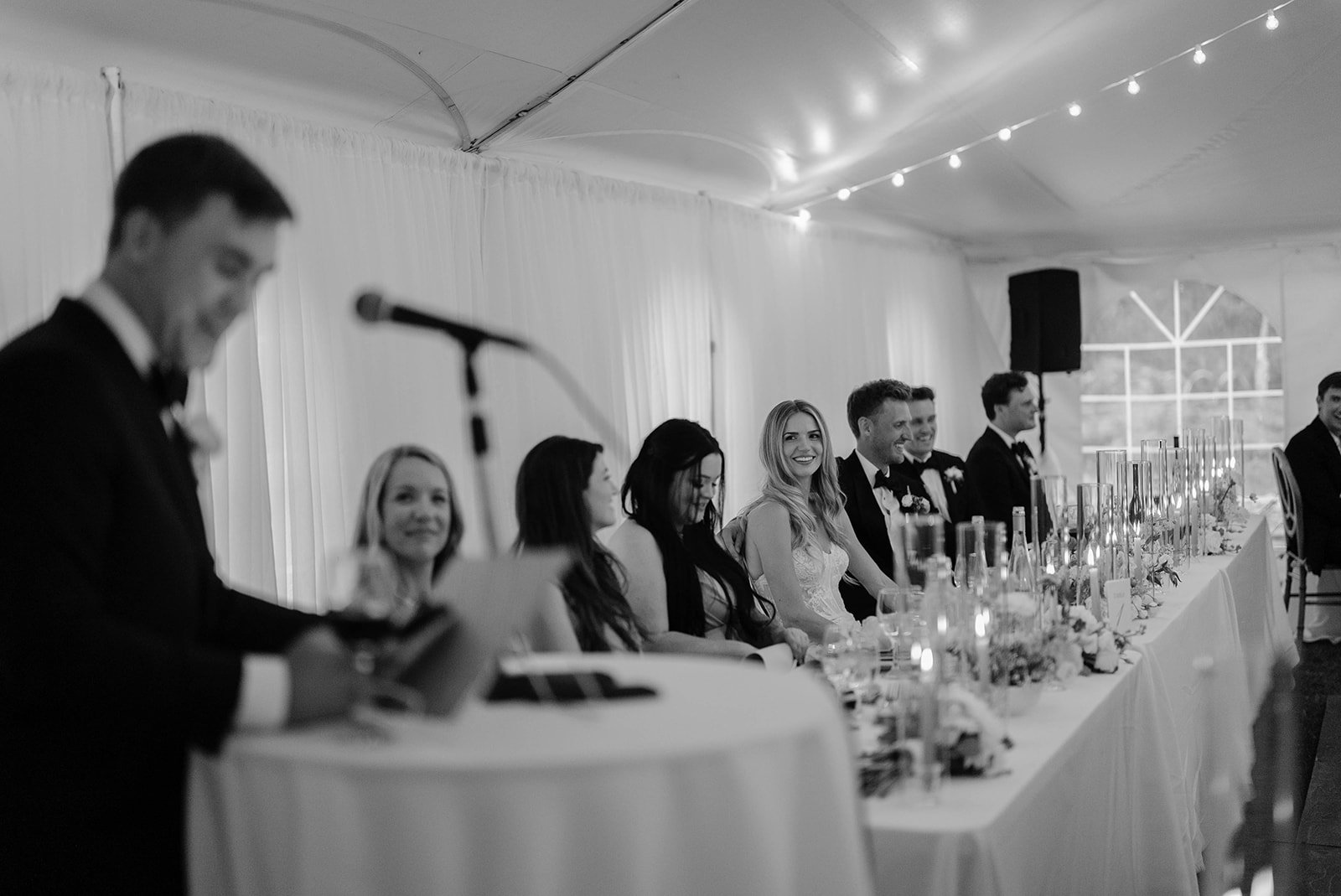 Black and white photo of a wedding reception with the bridal party seated at a long decorated table, and a man in a tuxedo giving a speech at a microphone.