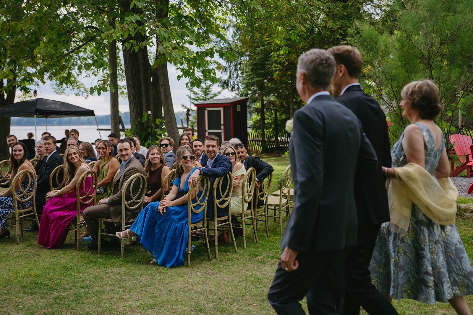 People attending an outdoor wedding ceremony, seated on gold chairs, with three individuals walking down the aisle, surrounded by trees and water in the background.