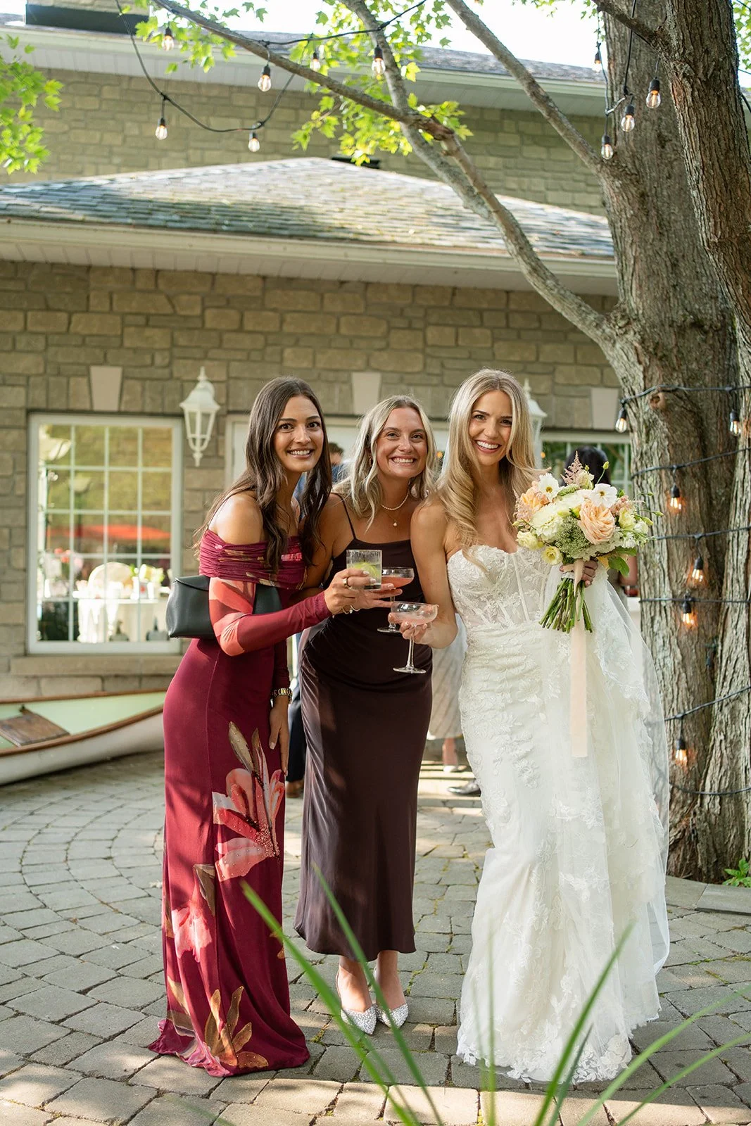 Three women standing outdoors at a celebration, with one in a white wedding dress holding a bouquet, in front of a house with string lights and a large tree.