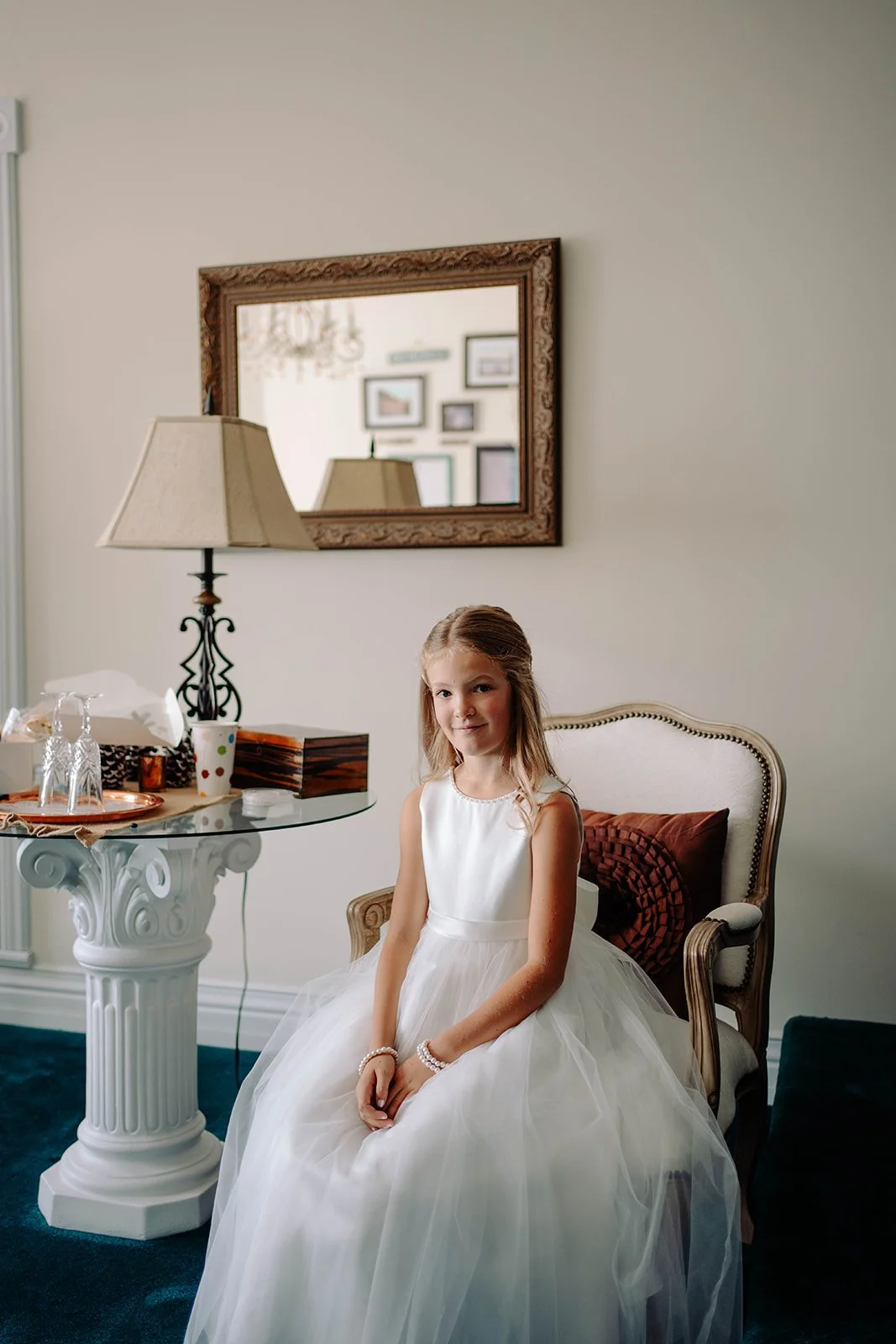 A young girl in a white dress sitting on an elegant armchair in a room, with a marble table with glasses and a lamp next to her, and a mirror and framed pictures on the wall behind her.