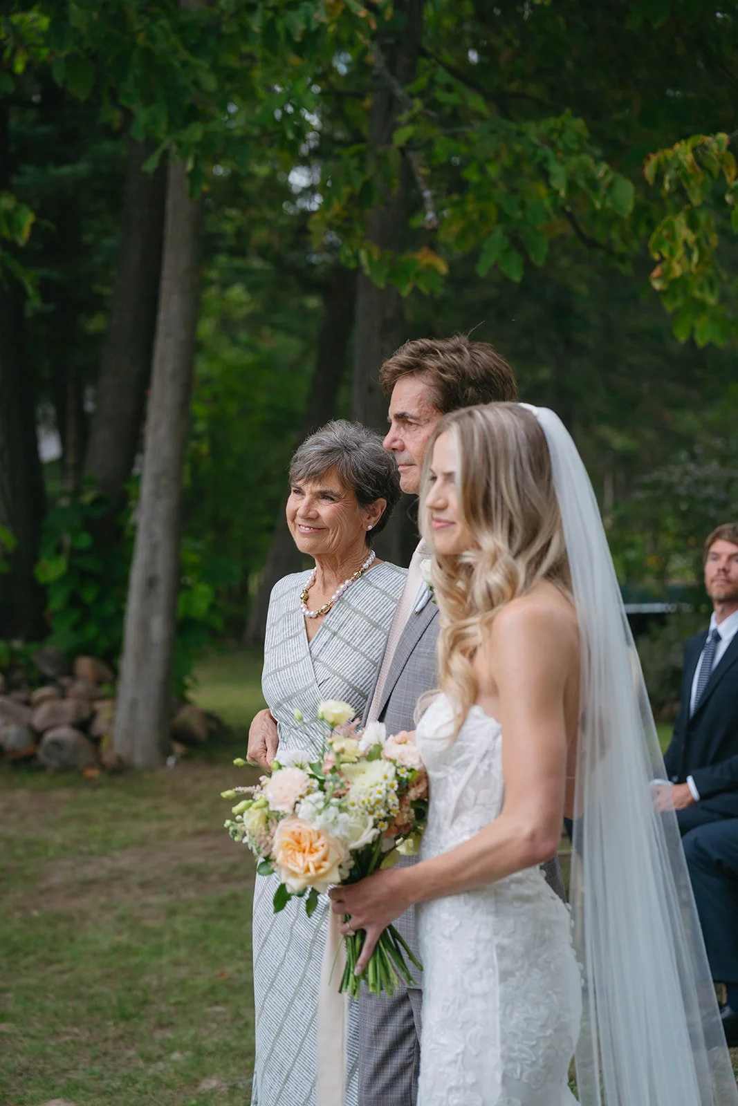 Bride in white wedding gown holding a bouquet, standing next to an older woman in a patterned dress and a man in a suit, outdoors with green trees in the background.