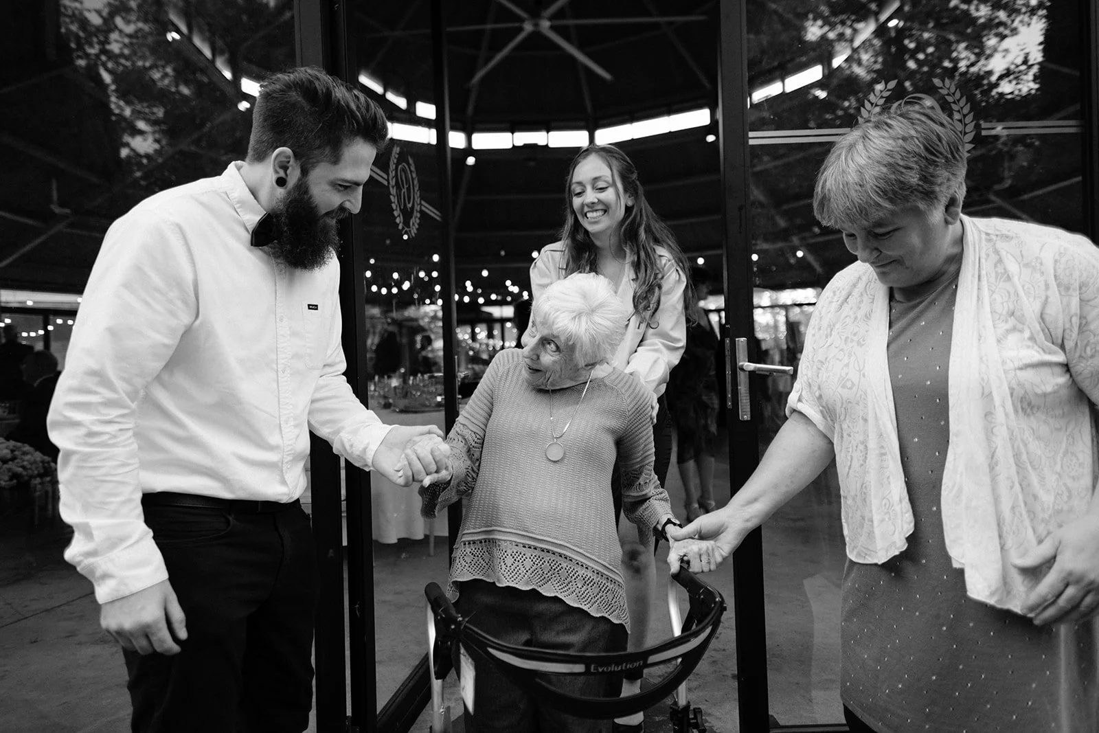 Four people, including an elderly woman in a wheelchair, are smiling and holding hands in a circle. They are inside a building with glass doors, celebrating and enjoying each other's company.