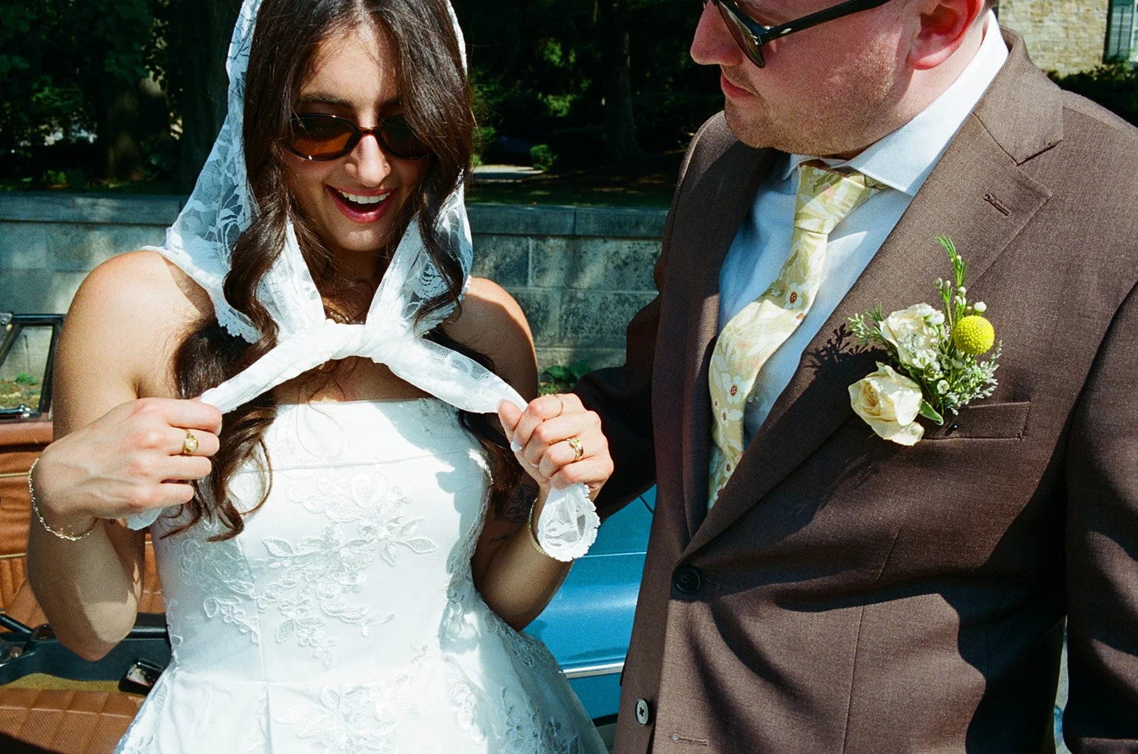 A woman in a white wedding dress and sunglasses smiling, holding a white lace scarf. A man in a brown suit with a boutonnière is standing beside her, looking at her.