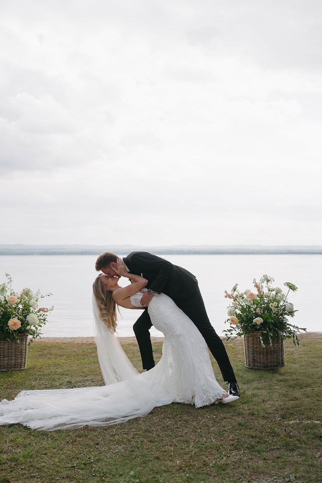 A bride and groom dancing outdoors near a lake, with large flower arrangements in baskets on either side.
