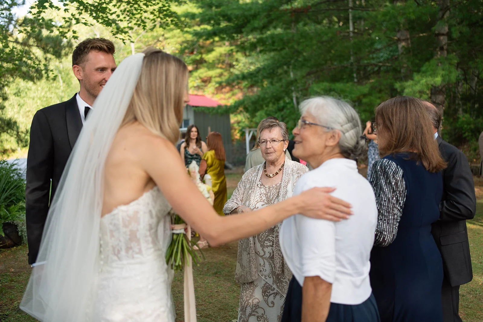 Bride in a white wedding dress with a veil talking to an older woman in a white blouse, while a groom in a tuxedo and other guests are nearby in a lush outdoor setting.