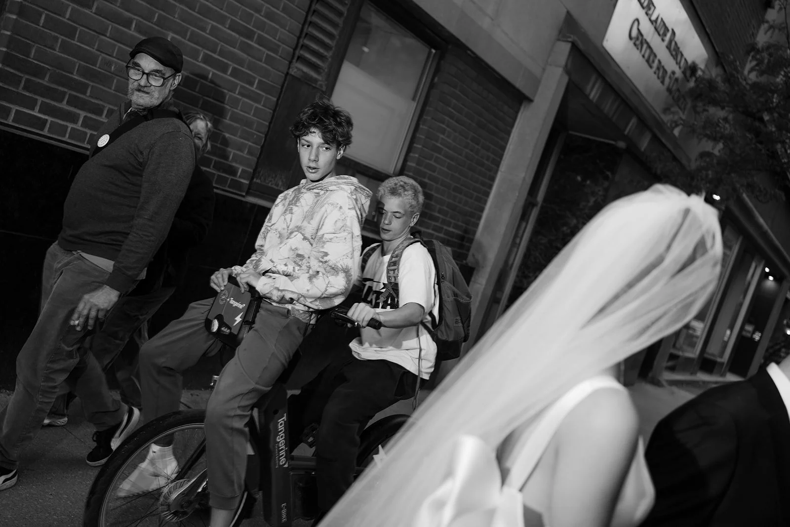 People walking on a city sidewalk at night, including a young man on an electric bike, a woman, and a man walking past them.