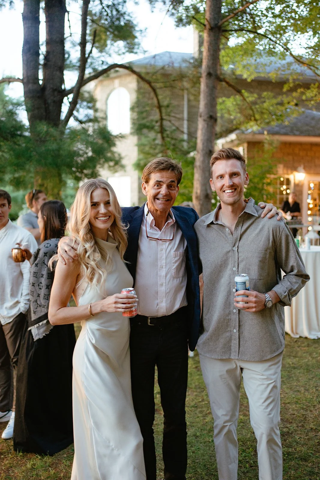 Three people standing outdoors at a social gathering, smiling, with drinks in hand. There are trees and a building in the background, and other guests are visible behind them.