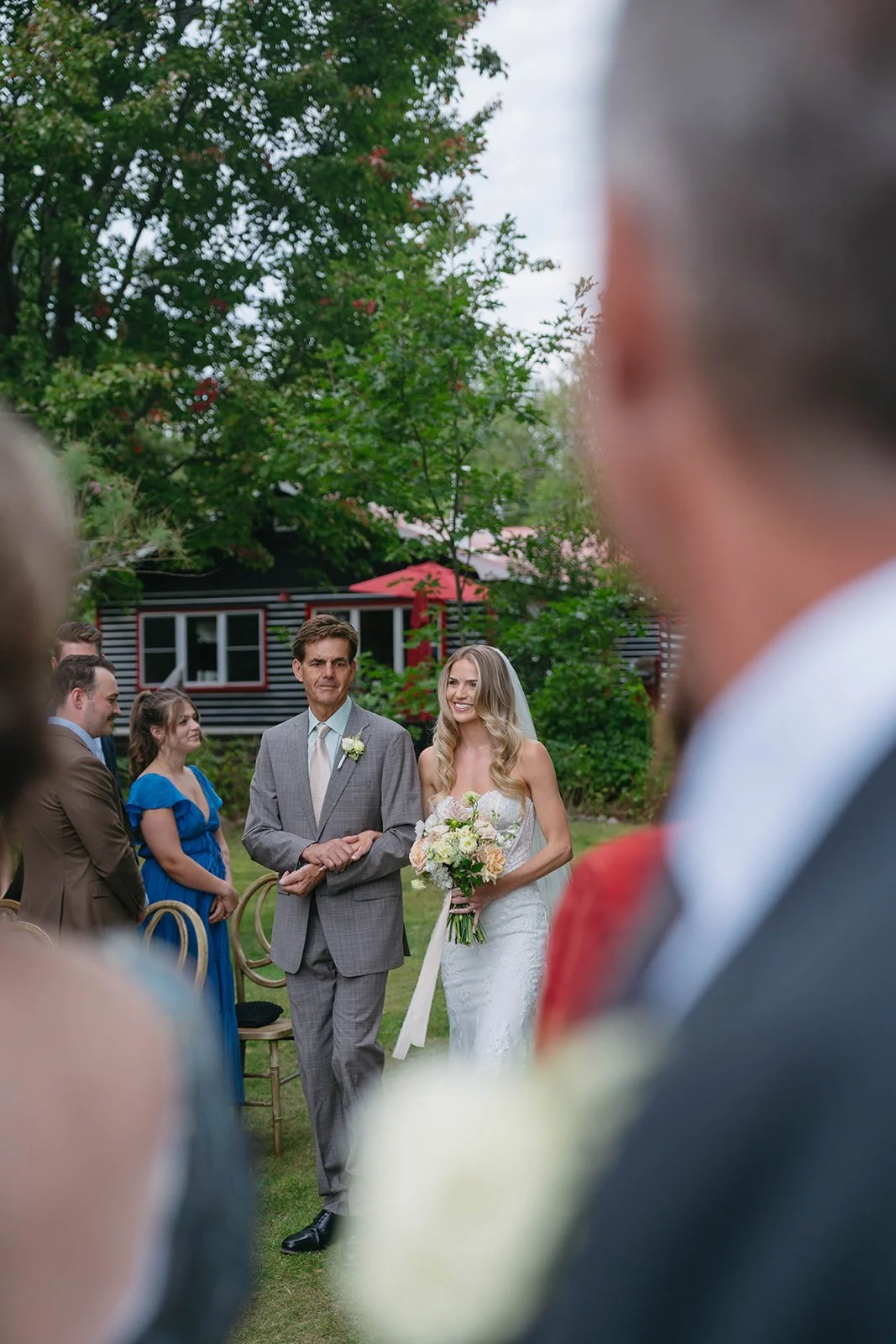 Bride walking down the aisle with her father during an outdoor wedding ceremony, with guests seated on either side and trees in the background.